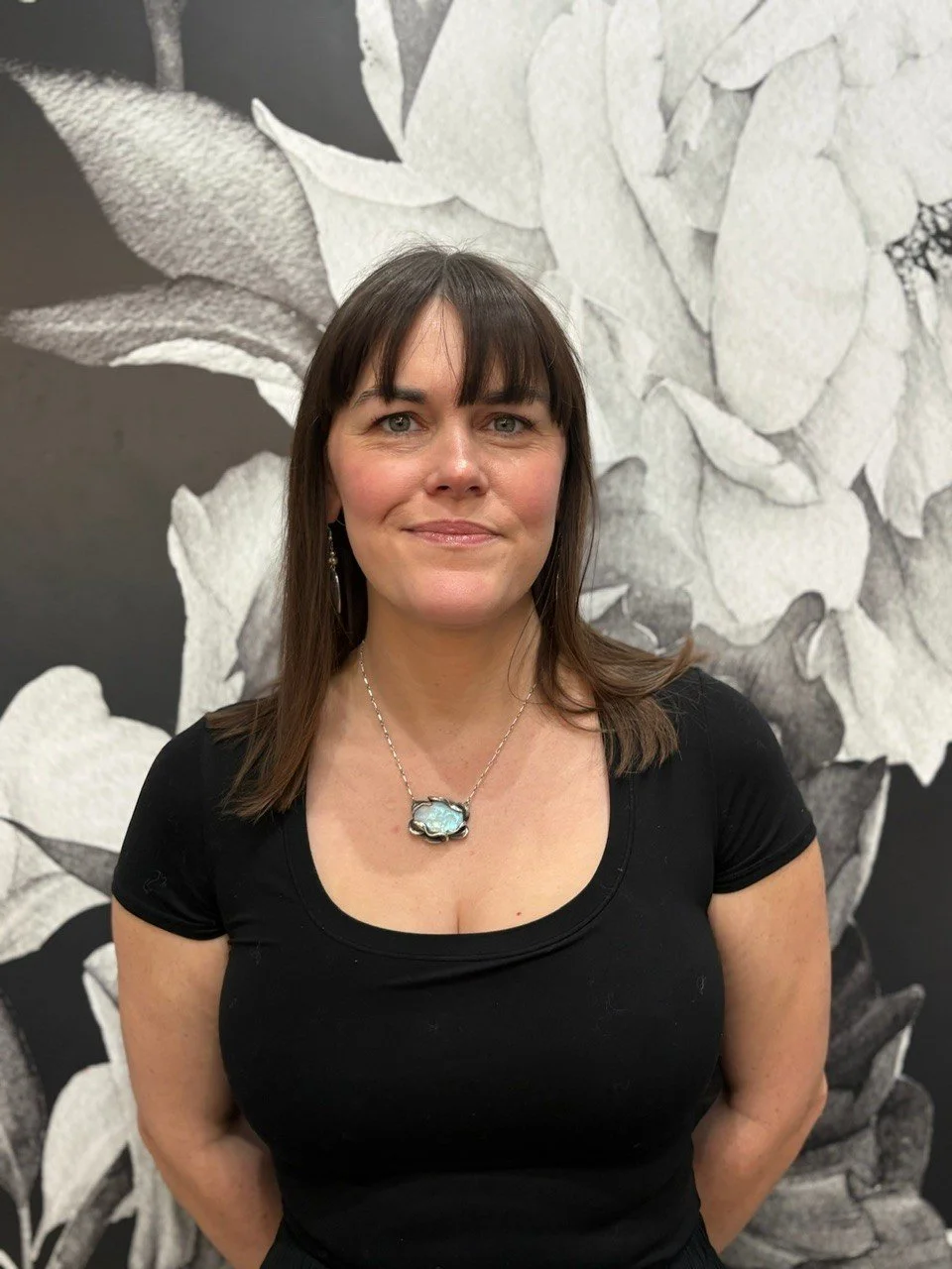 A woman with shoulder-length brown hair and bangs standing in front of a black and white floral mural. She is wearing a black shirt and a silver necklace with a large oval-shaped pendant.
