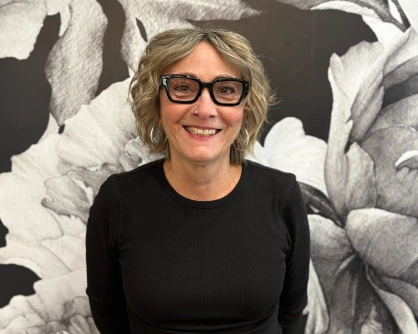 A woman with curly blonde hair and black glasses smiling, wearing a black top, standing in front of a black and white floral mural.