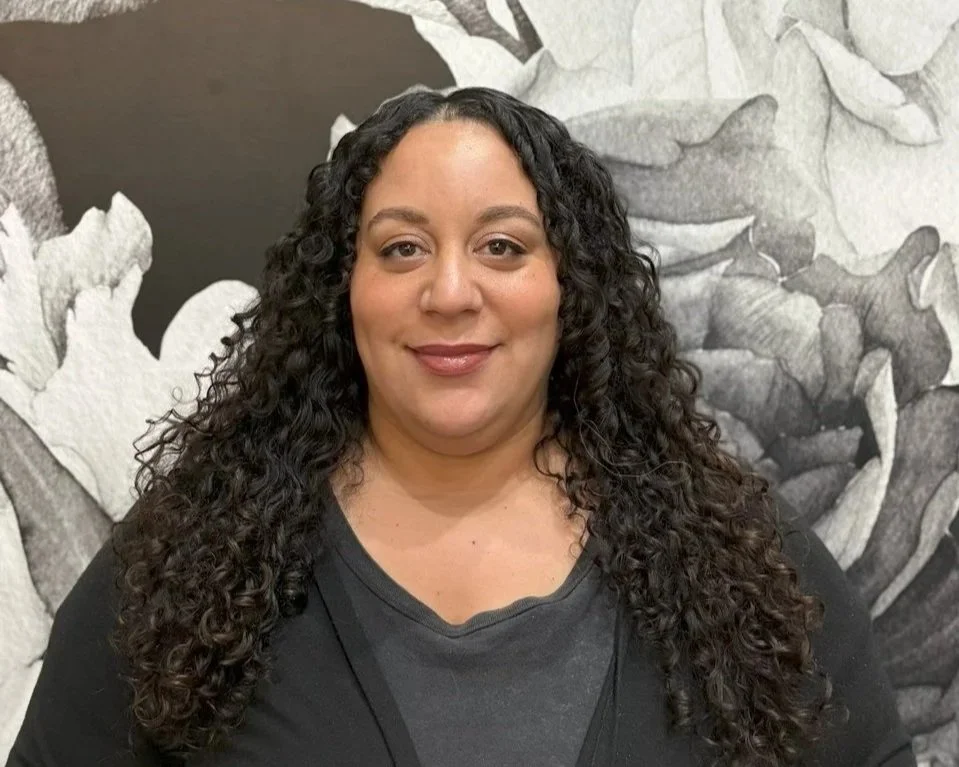 A woman with long, curly black hair and a black shirt stands in front of a black and white floral mural.