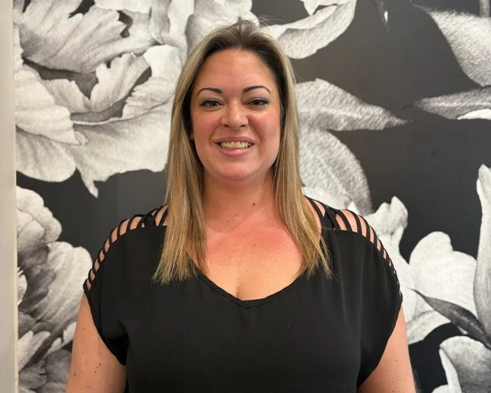 A woman with blonde hair smiling, wearing a black top with cut-out shoulder details, standing in front of a black and white floral wallpaper background.