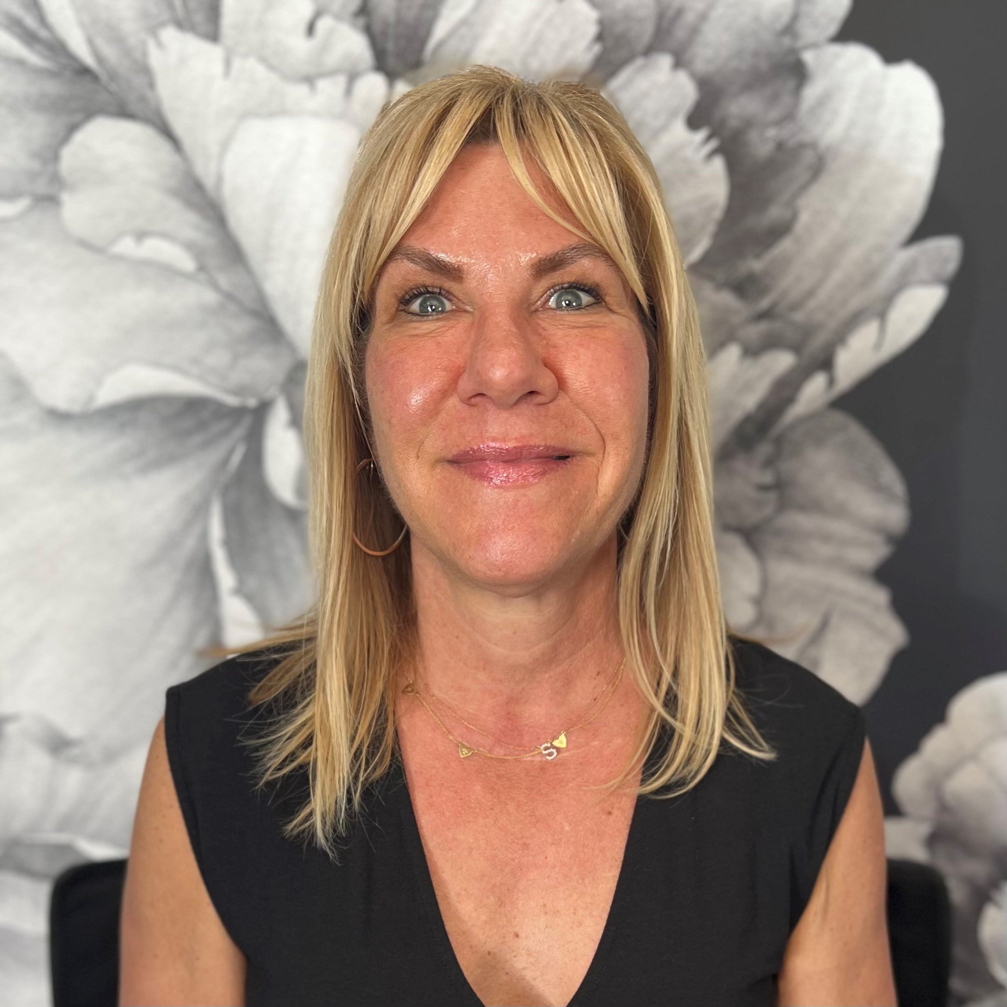 A woman with shoulder-length blonde hair, wearing a black sleeveless top, gold hoop earrings, and layered gold necklaces, smiling in front of a grayscale floral background.