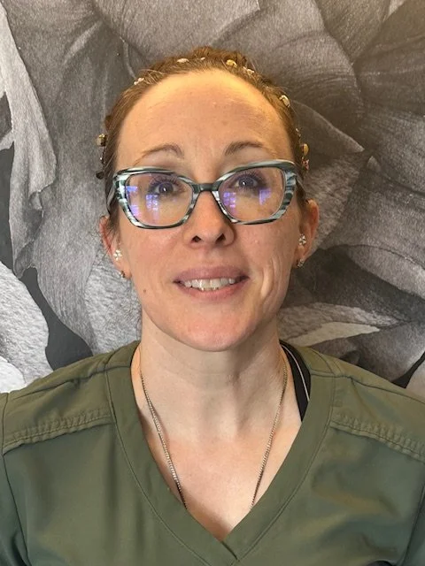 A woman with glasses, red hair, and a green top smiling for the camera, standing in front of a black and white mural of a dog's face.