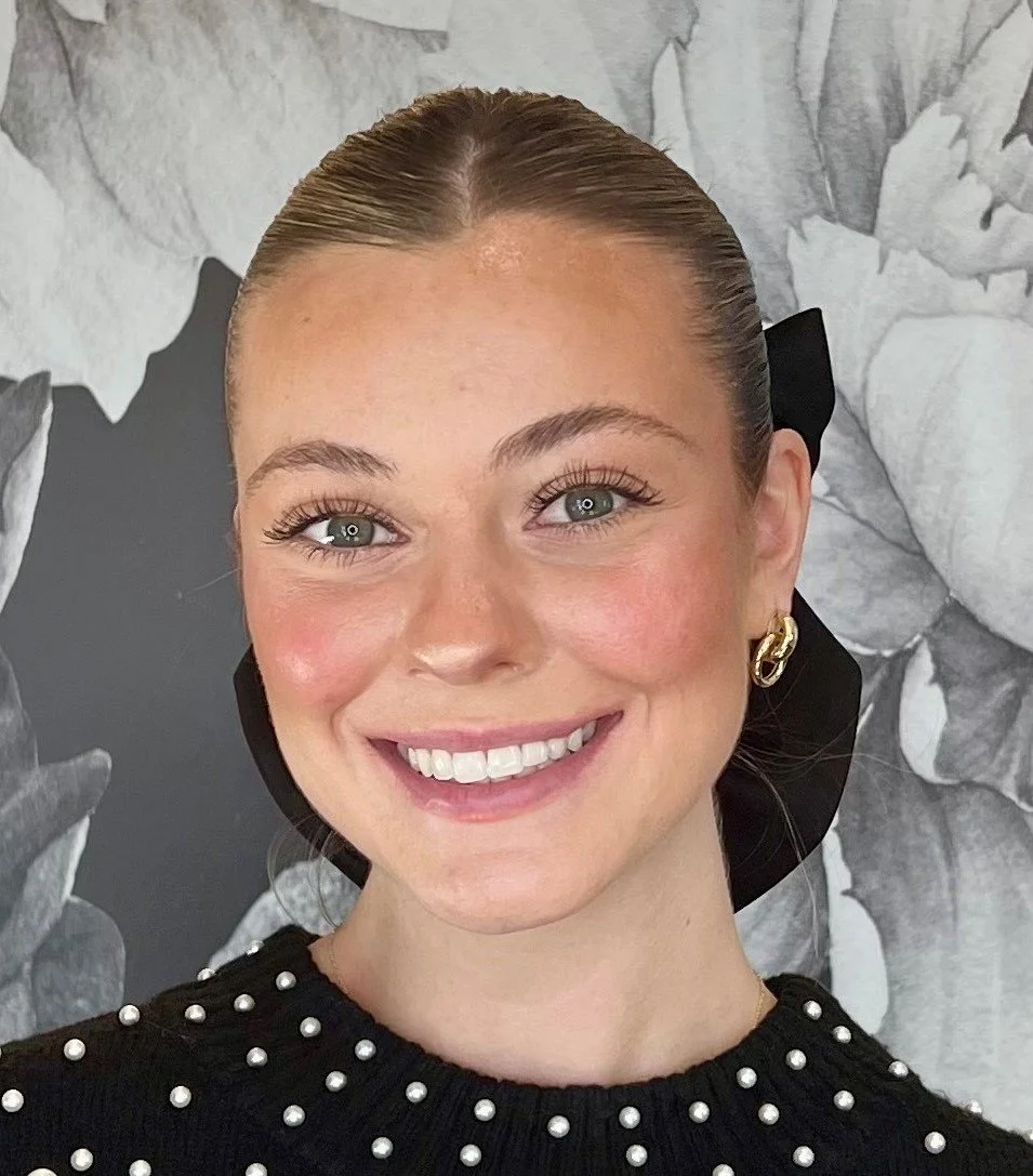 A young woman with blonde hair tied back, wearing a black sweater with pearl embellishments and gold hoop earrings, smiling in front of a black and white floral background.