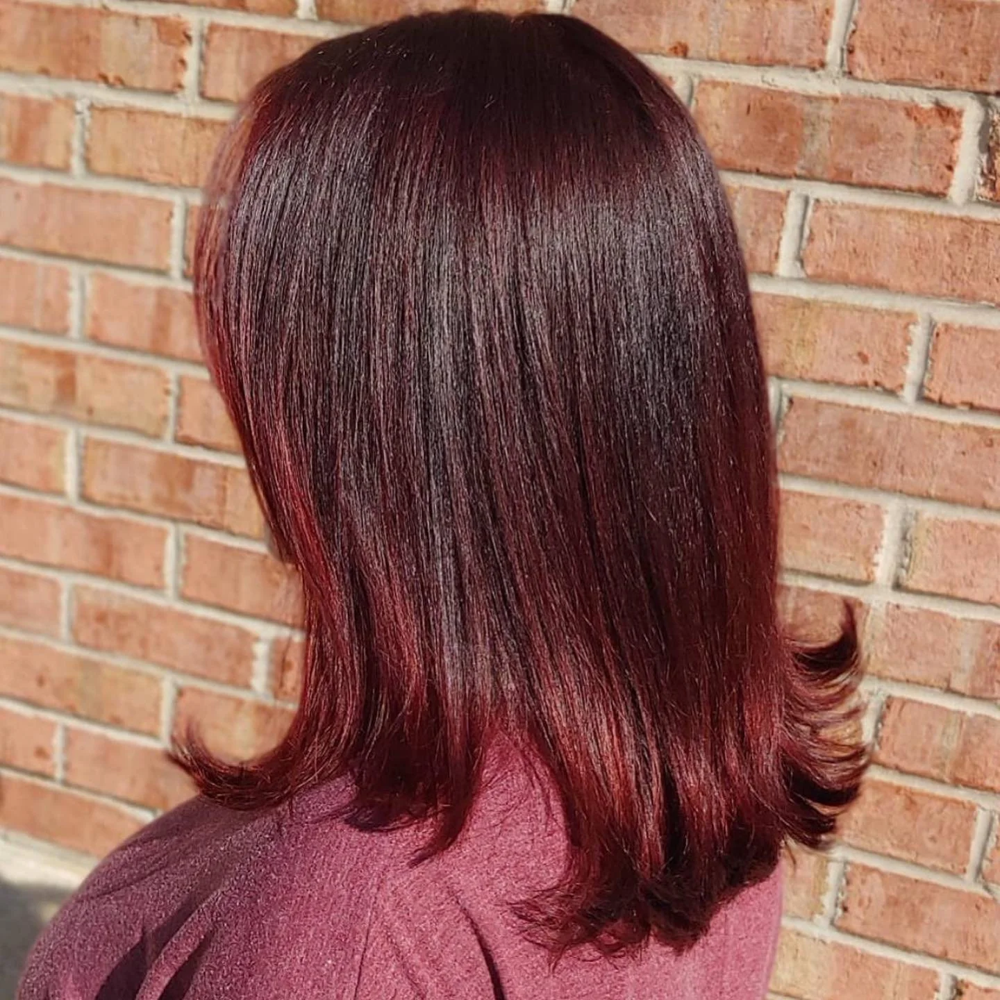 Woman with shoulder-length, shiny, dark red hair standing in front of a brick wall.