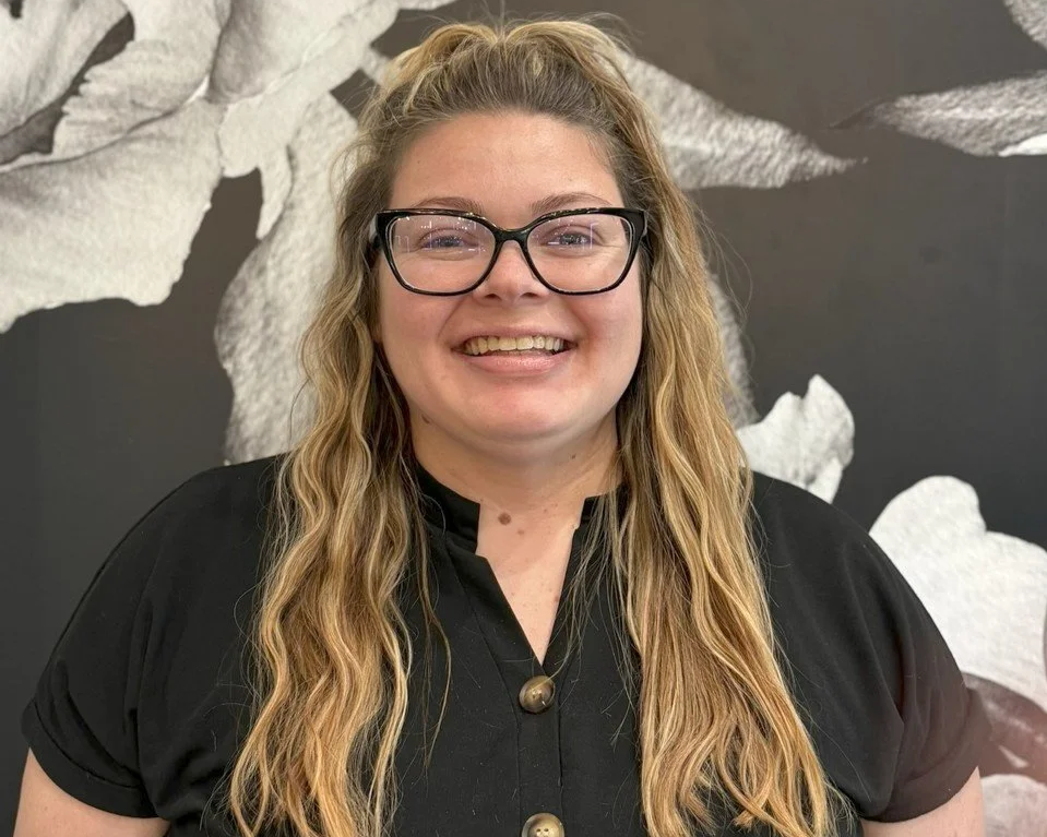 A woman with long blonde hair, glasses, and wearing a black top, smiling in front of a black and white floral mural.
