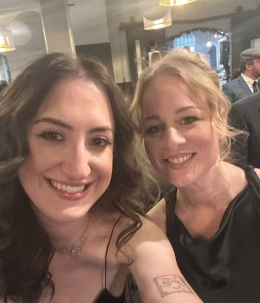 Two women smiling at a restaurant or café, with other people in the background.