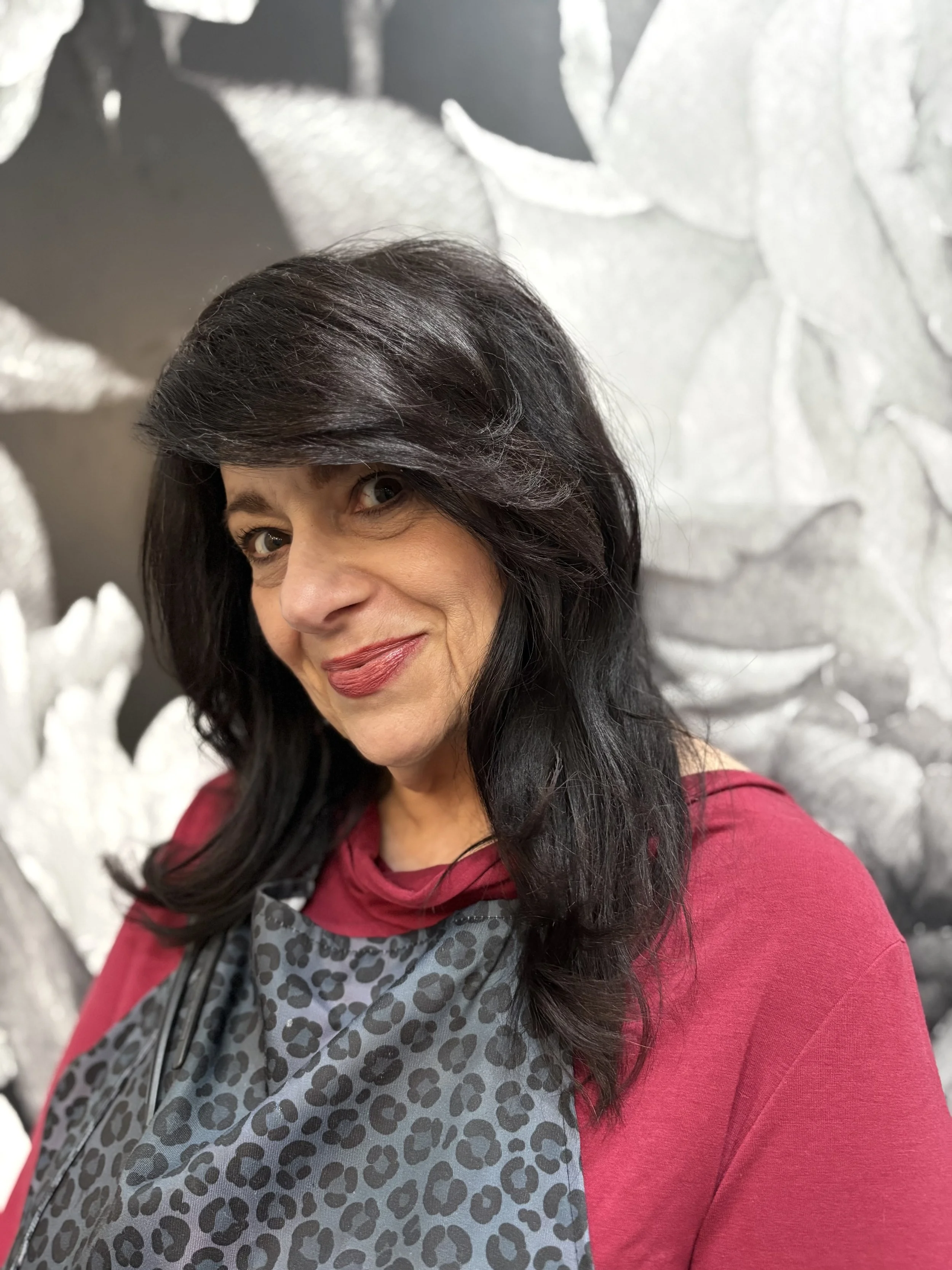 Woman with dark hair and red lipstick taking a selfie against a black and white floral background in a salon apron.