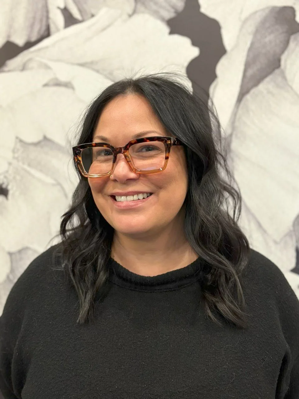 A woman with black wavy hair and tortoiseshell glasses smiling in front of a floral wall background.