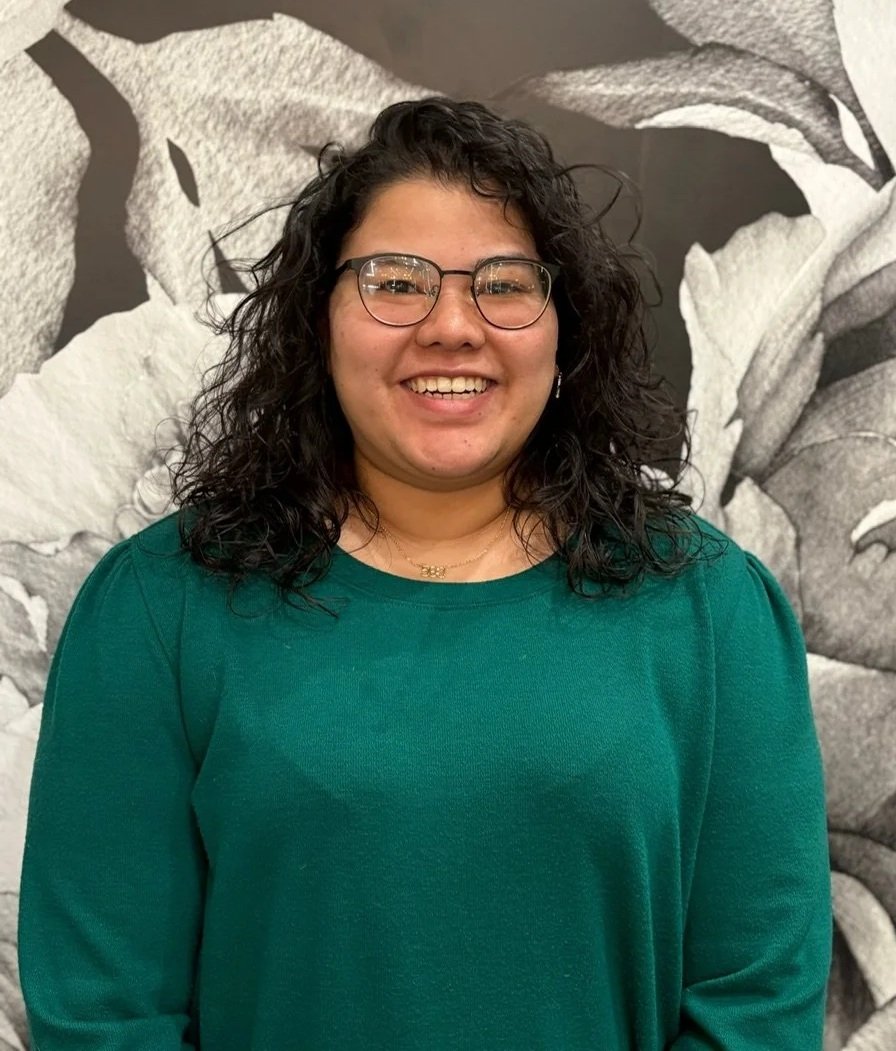 A woman with curly dark hair, glasses, and a wide smile, wearing a green sweater, standing in front of a large, black-and-white floral mural.