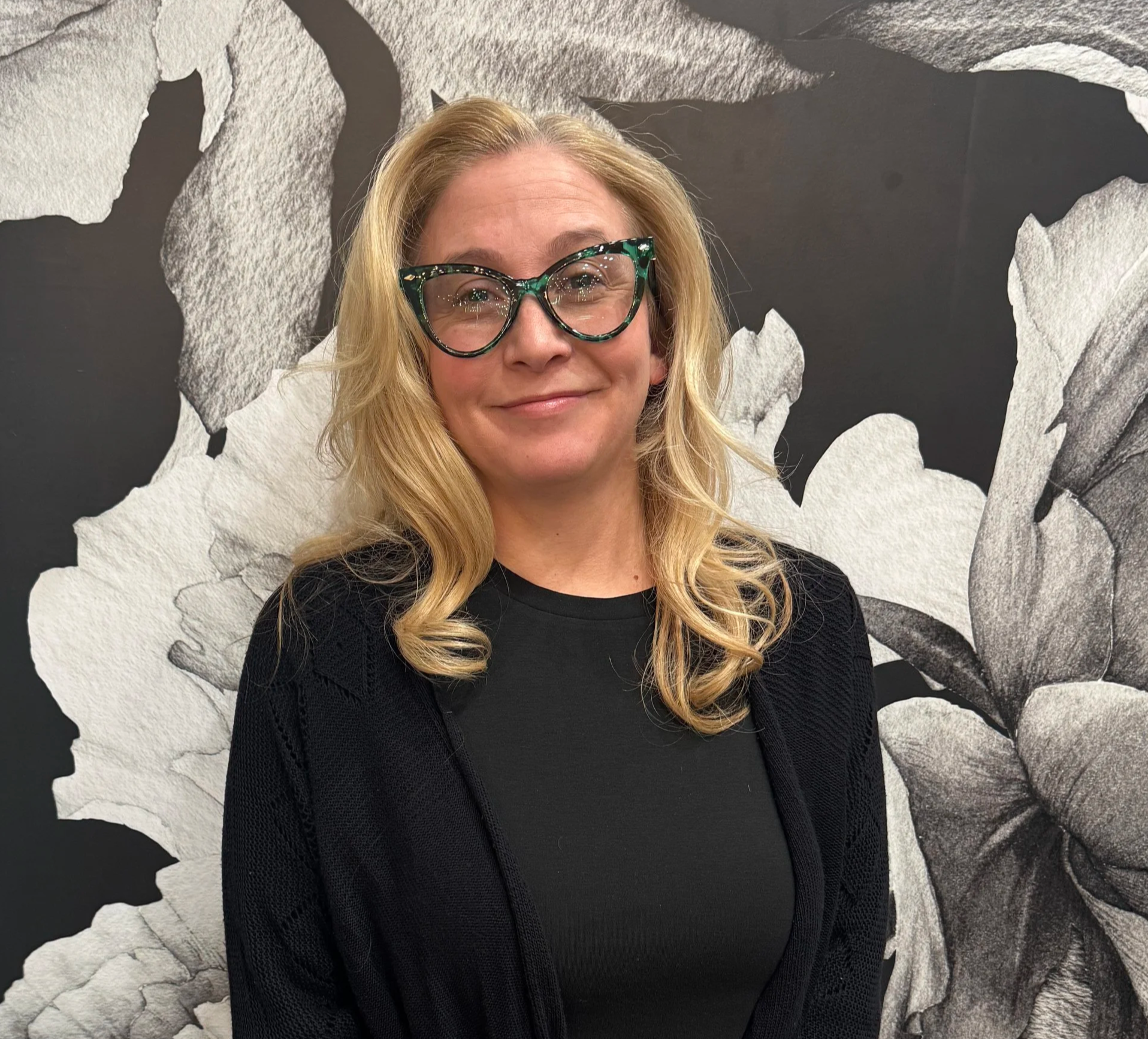 A woman with blonde hair, wearing glasses and a black top, smiling in front of a black and white floral patterned wall.