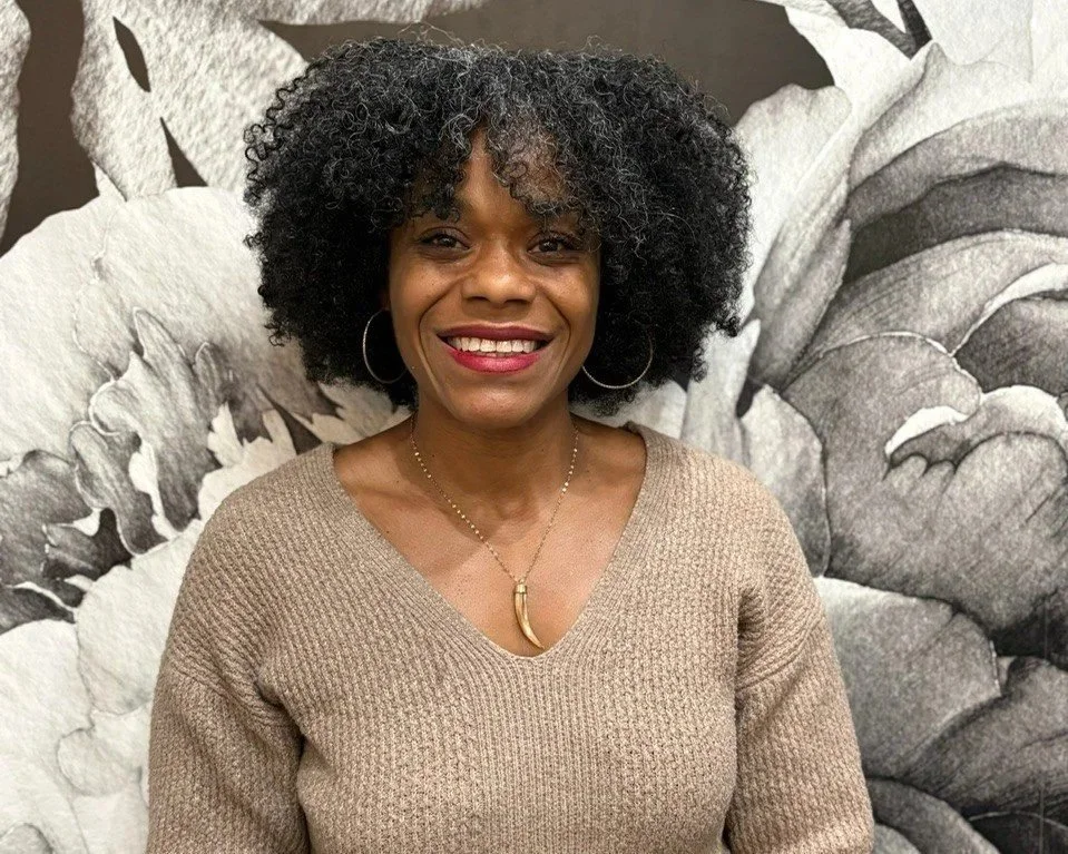 A woman with curly black hair and hoop earrings smiling in front of a black-and-white floral mural, wearing a beige sweater and a gold necklace.