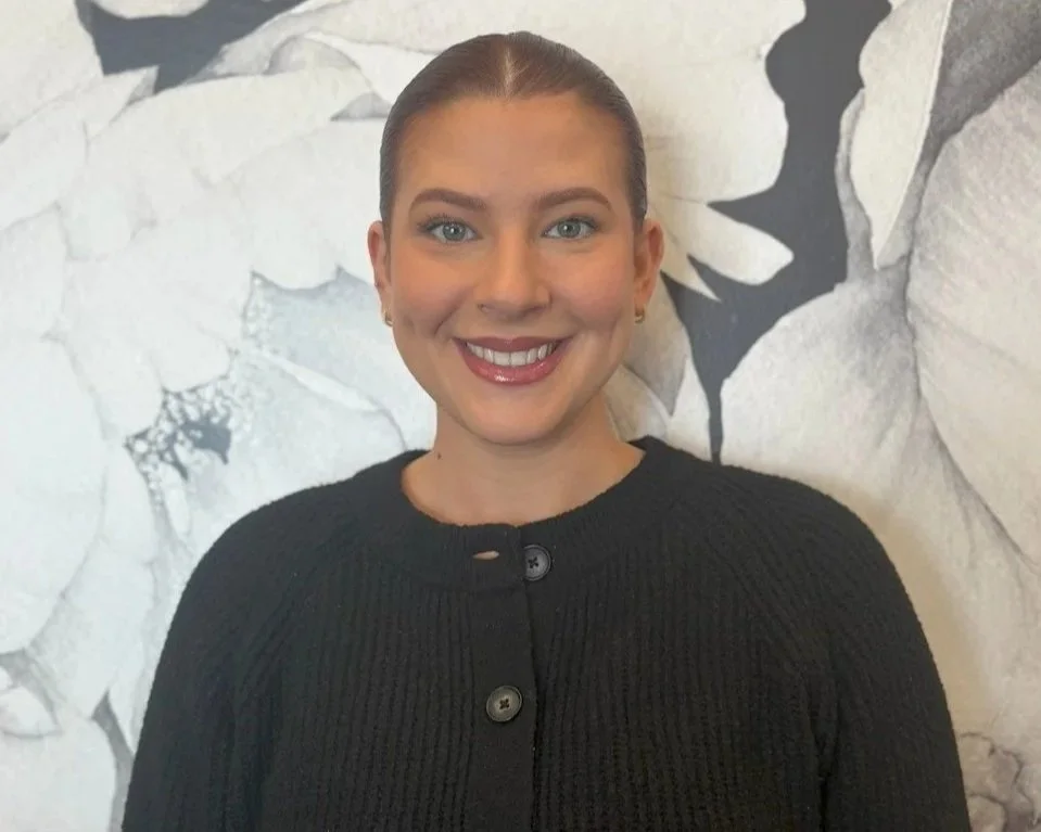 A woman with light skin, slicked back hair, wearing a black sweater, smiling in front of a white and black floral wallpaper background.