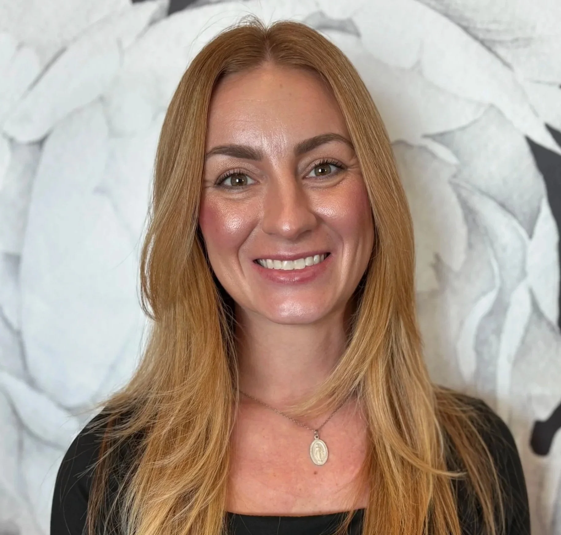 A woman with long red hair smiling, wearing a black top and a necklace with a white pendant, standing in front of a light-colored, textured background.