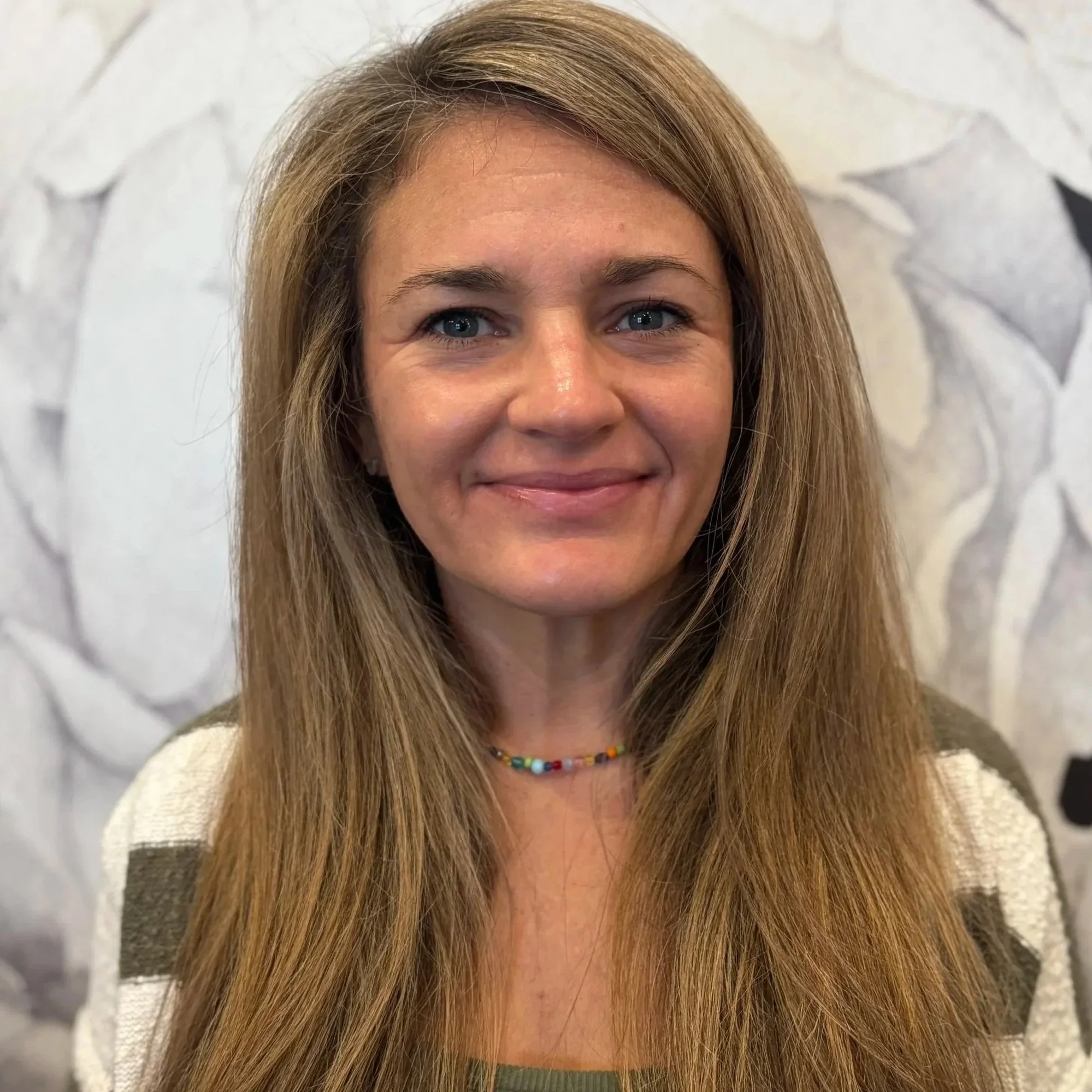 A woman with long, light brown hair and blue eyes smiling in front of a floral-patterned background.
