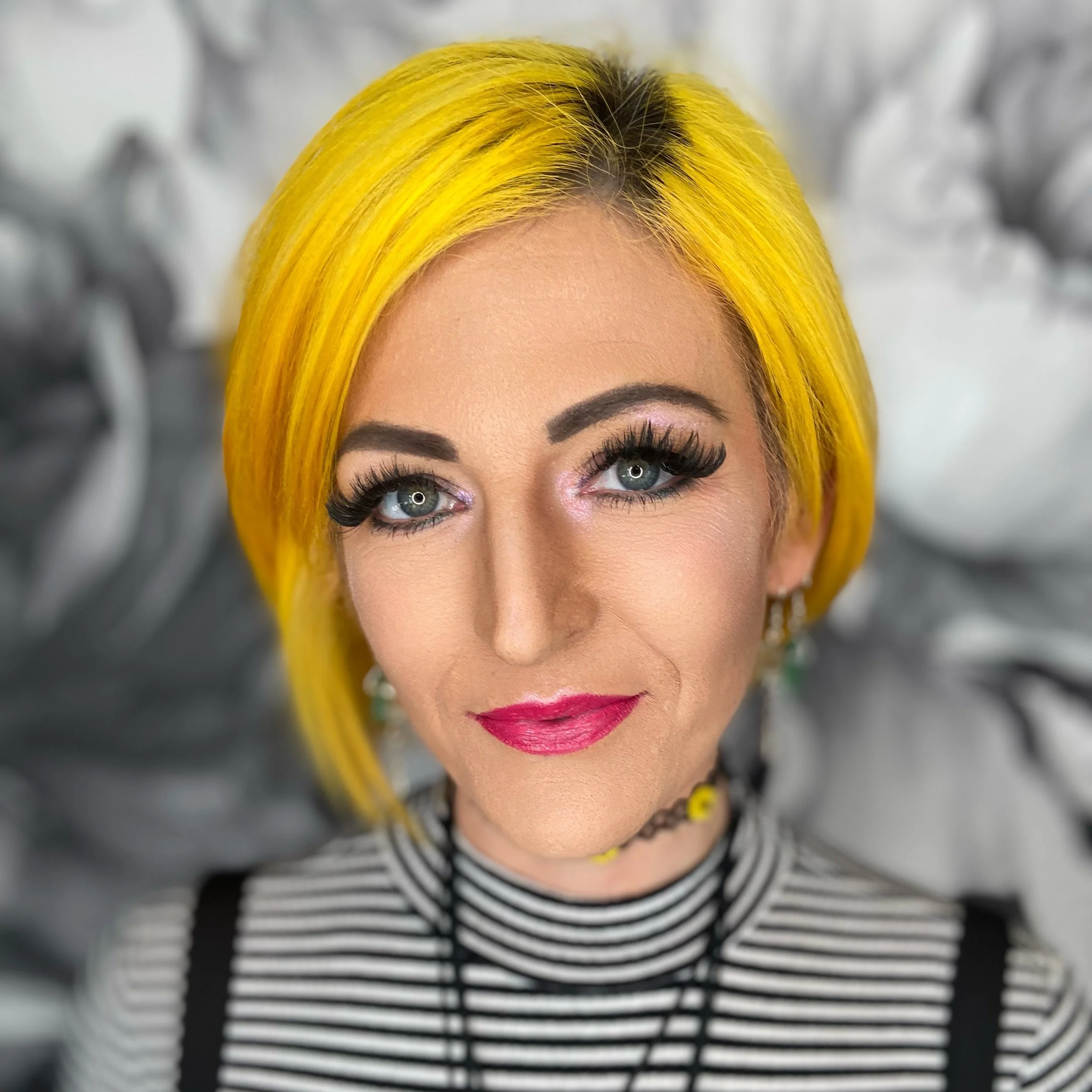 A woman with bright yellow hair, makeup, and a striped top. A blurred background features a tree or plant.