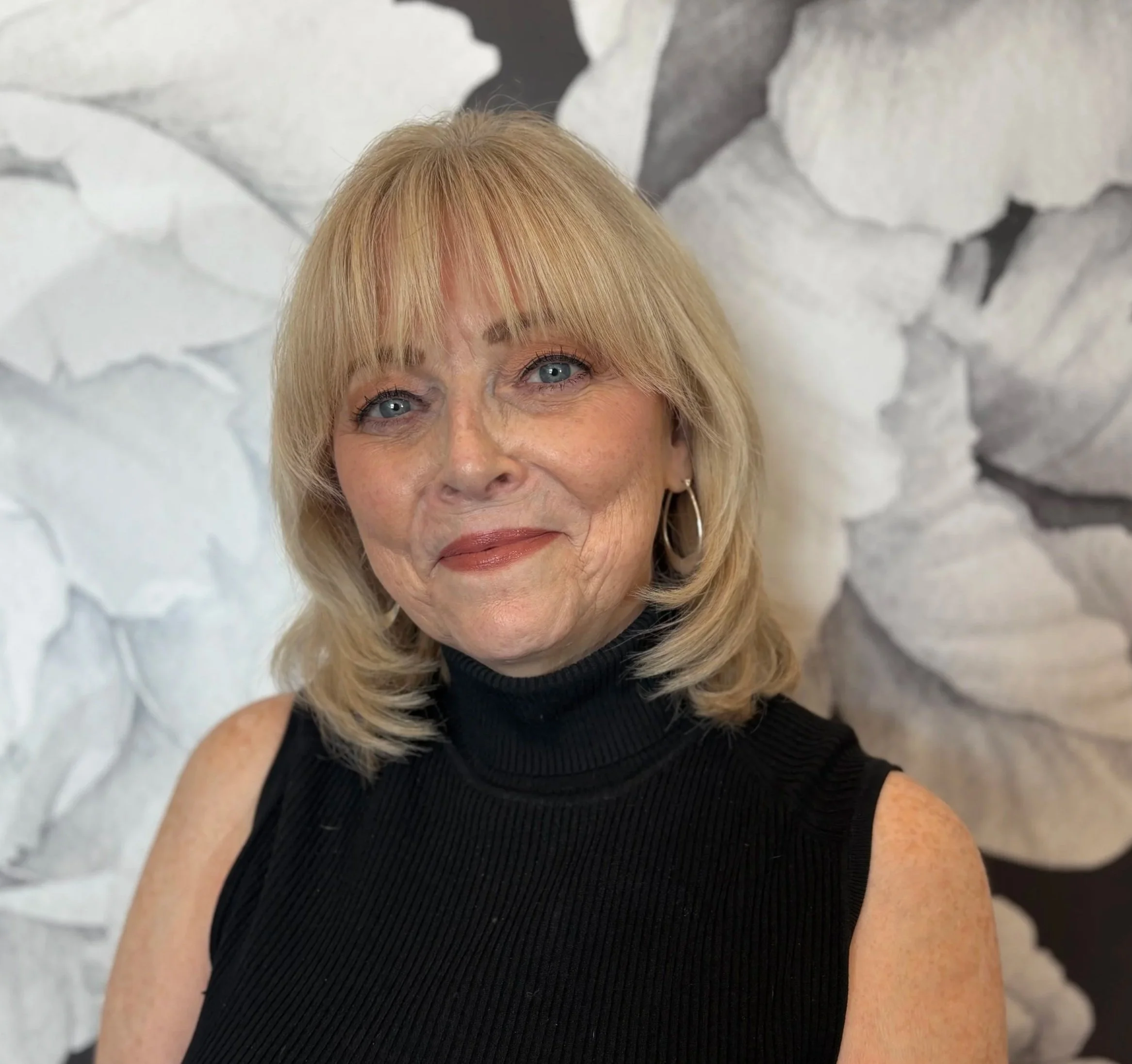 A woman with blonde hair, blue eyes, and hoop earrings, wearing a black sleeveless turtleneck, smiling in front of a textured black and white floral background.