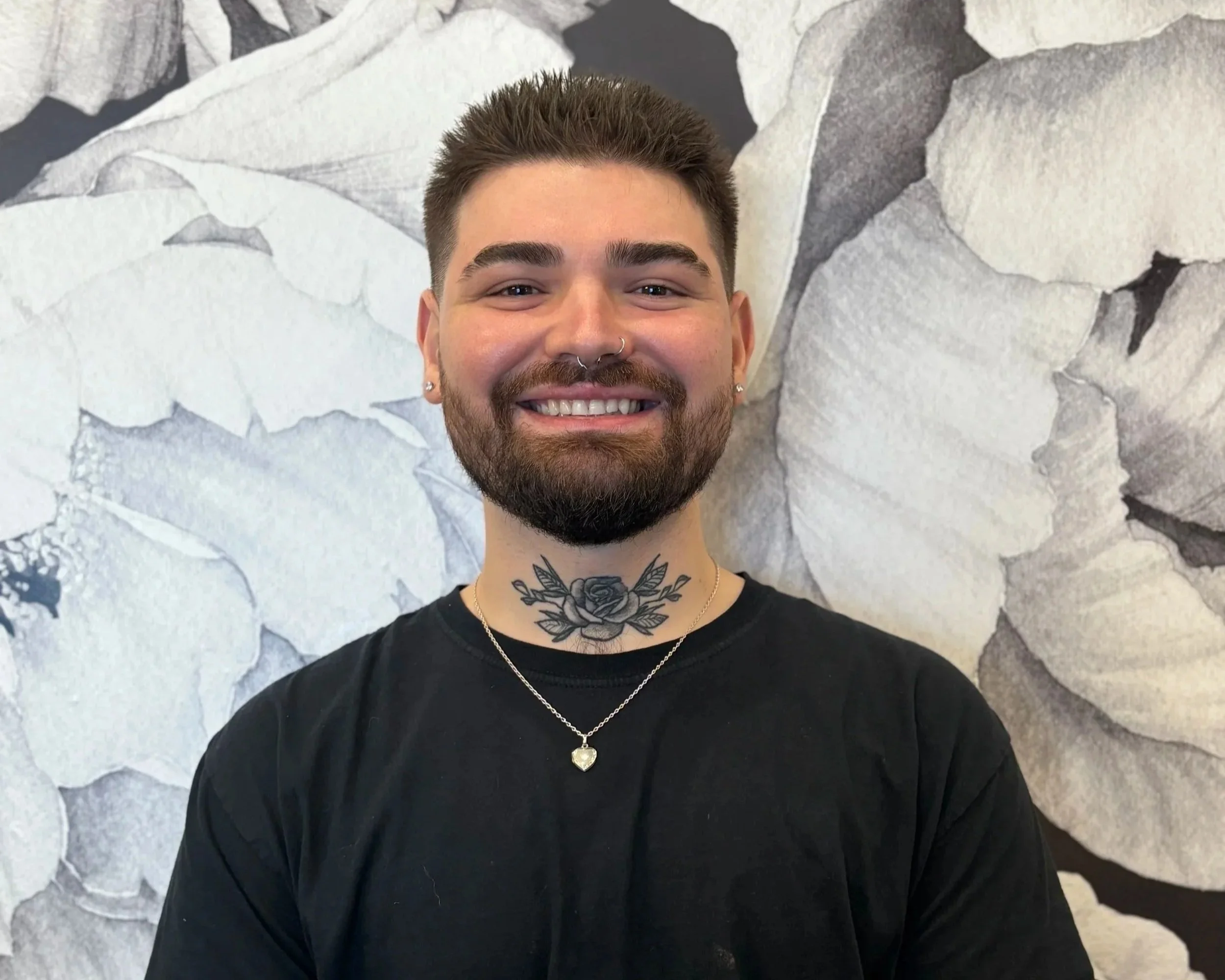 A young man with short dark hair, a beard, and a septum piercing, smiling at the camera, wearing a black shirt and a silver necklace with a heart pendant, standing in front of a large, abstract leaf mural in grayscale.