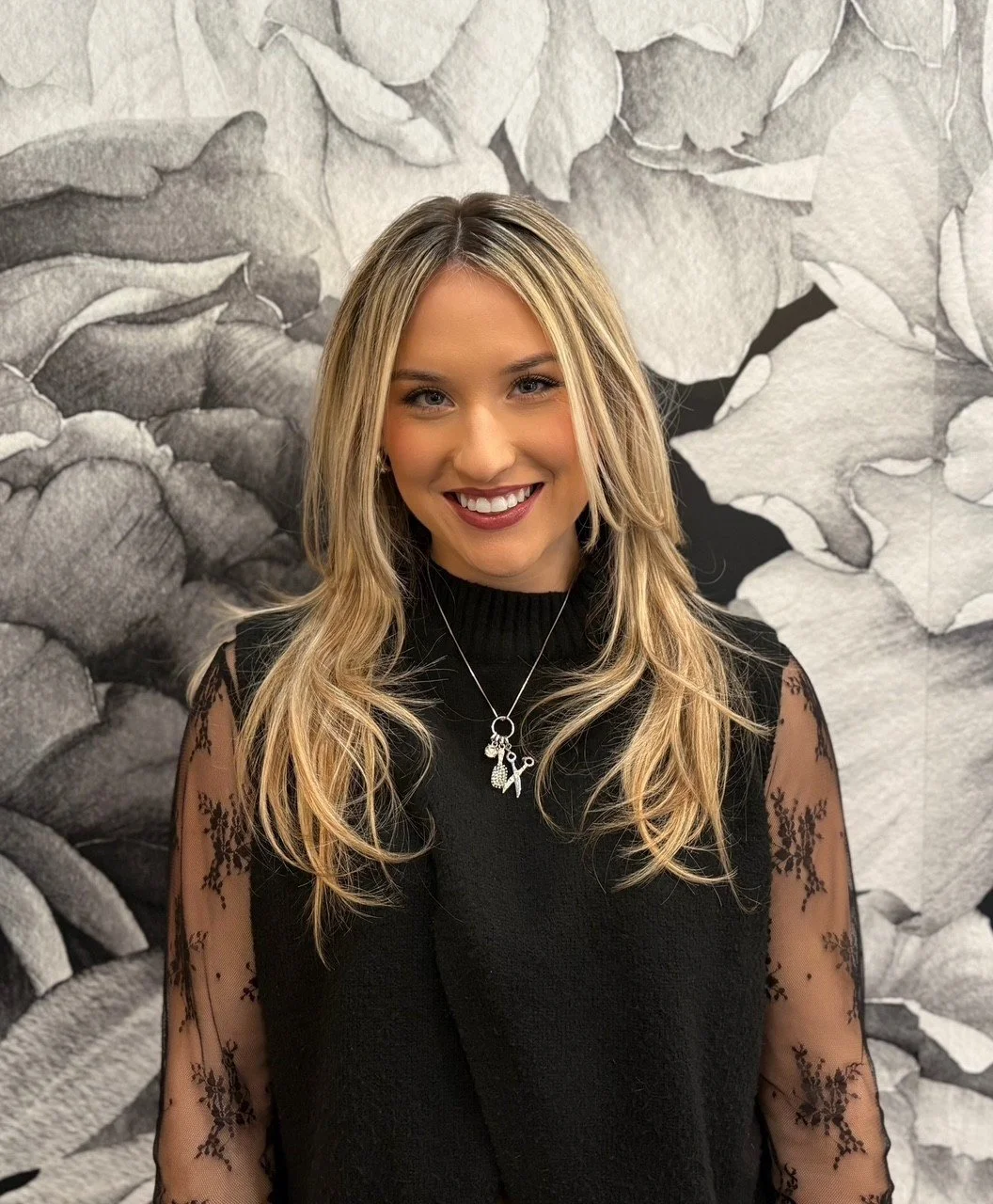 A woman with blonde hair smiling, wearing a black top with sheer, embroidered sleeves, and a necklace with graduation-themed charms, standing in front of a black and white floral mural.