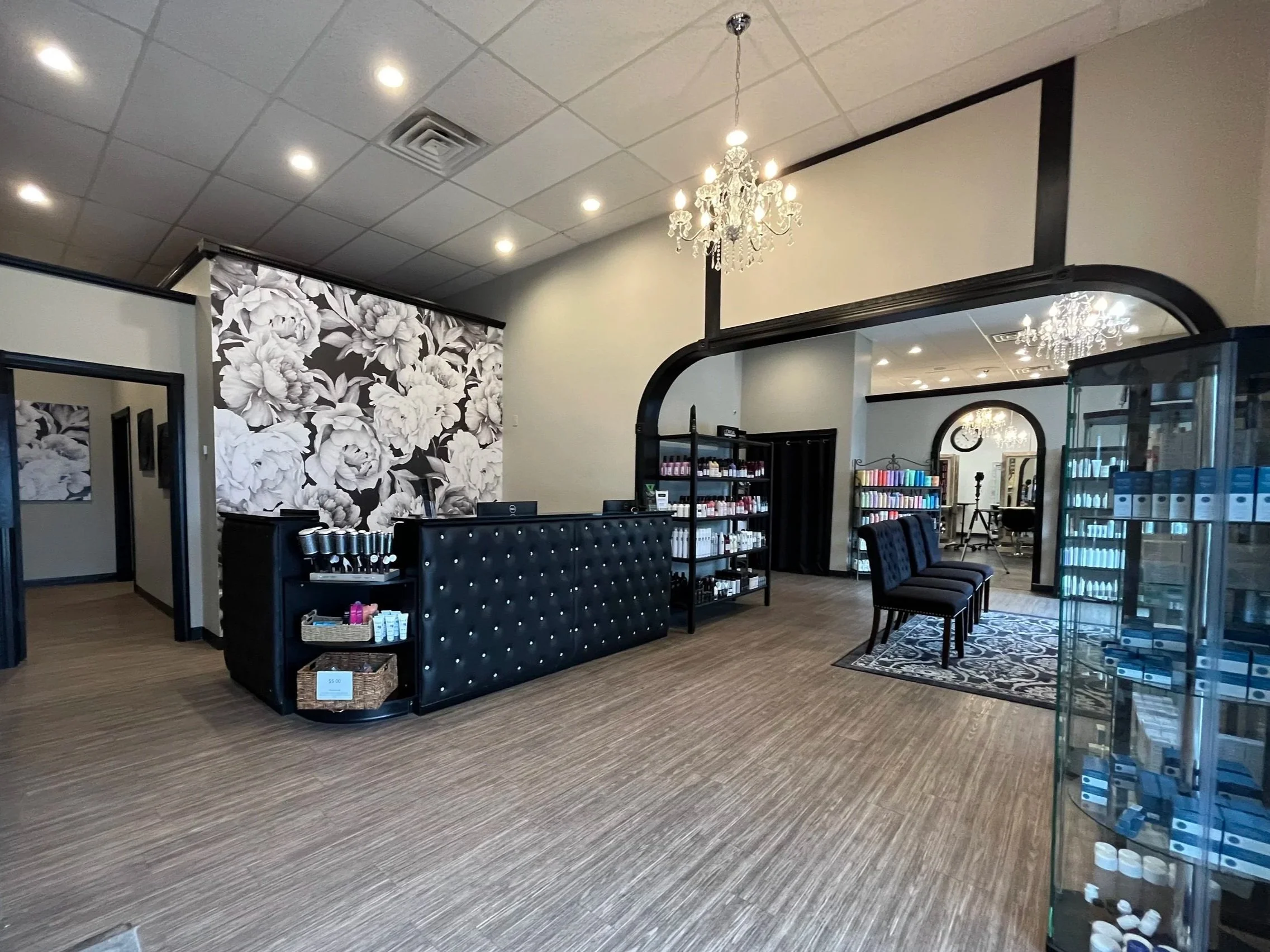 Inside our salon and spa with a reception desk, black chairs, shelves with products, a large floral black-and-white wall, a chandelier, and a mirror in the background.