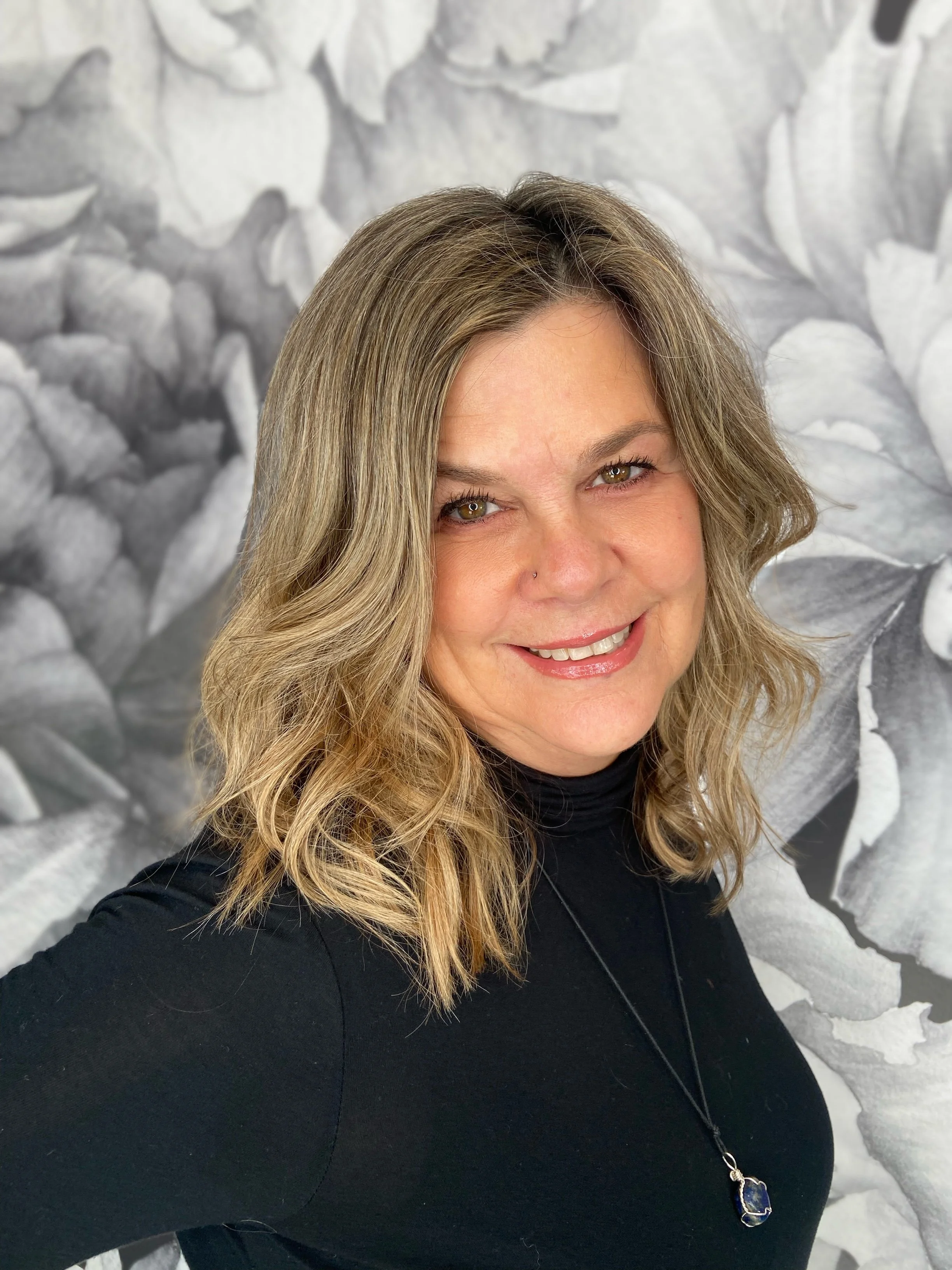 A woman with wavy, shoulder-length blonde hair smiling, wearing a black top and a necklace with a large pendant, standing in front of a large black and white floral background.