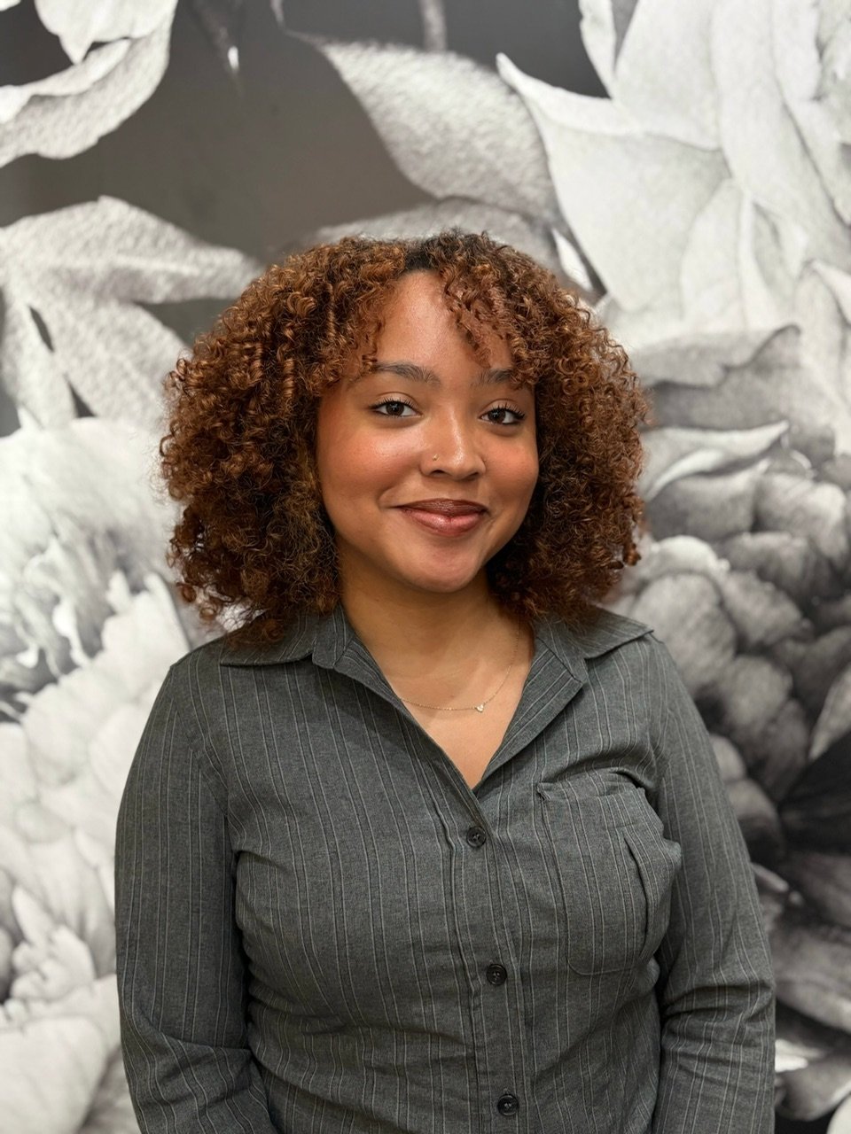 A woman with curly, reddish-brown hair smiling in front of a leafy black and white background.