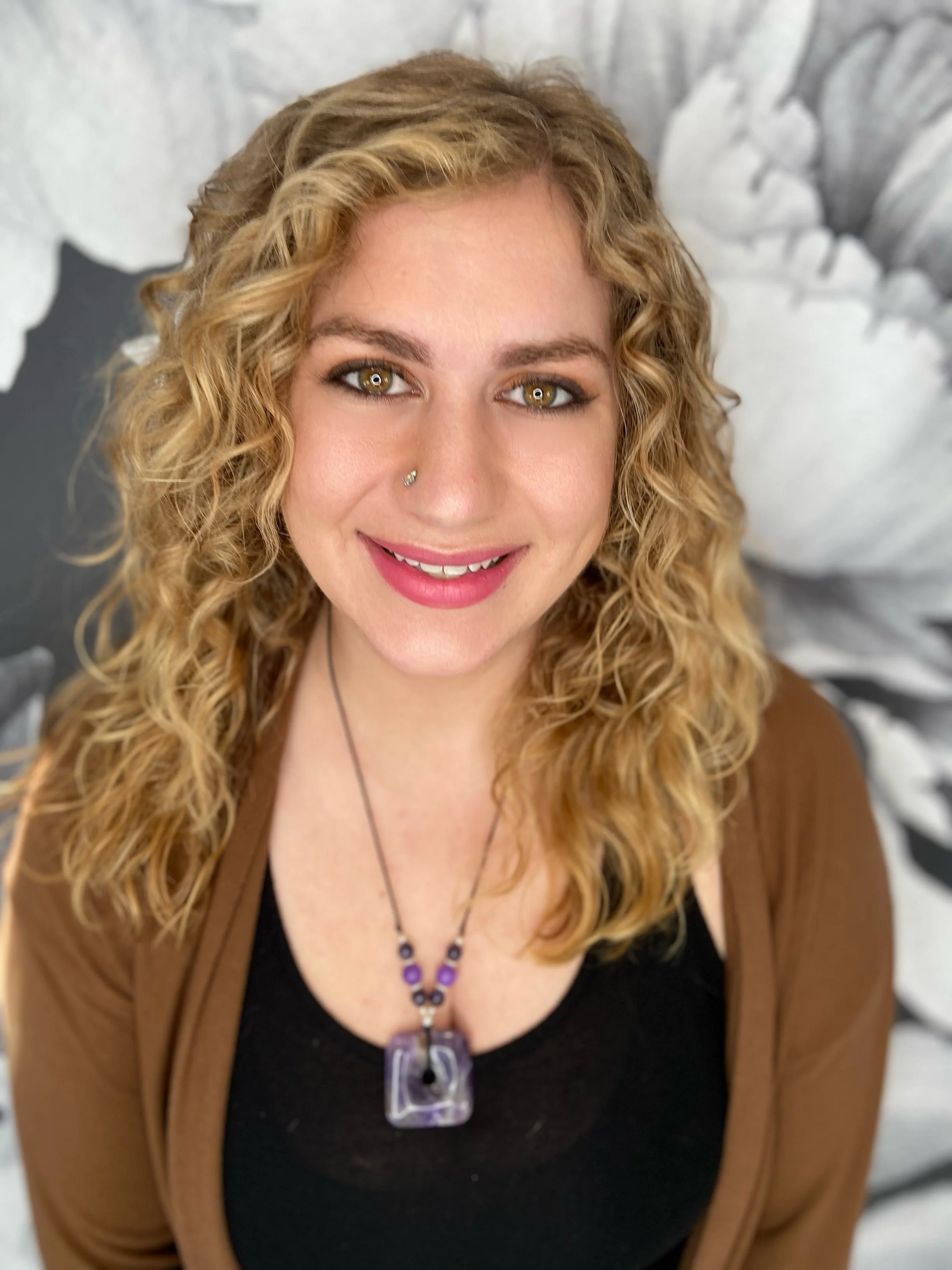 A woman with wavy blonde hair, hazel eyes, and a nose piercing, smiling in front of a black and white floral background. She is wearing a brown cardigan, a black top, and a purple pendant necklace.