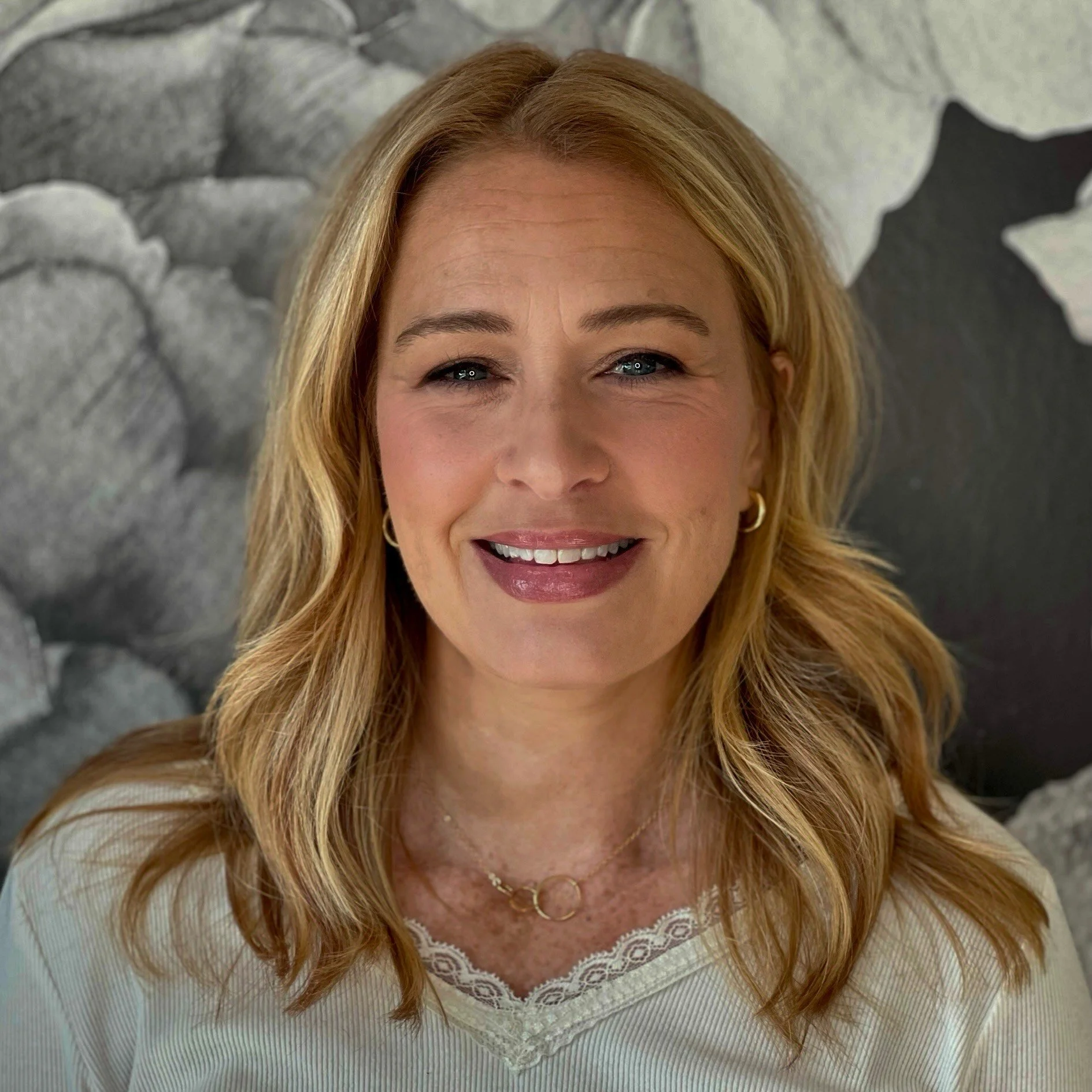 A woman with blonde, wavy hair and blue eyes, smiling in front of a stone wall background.