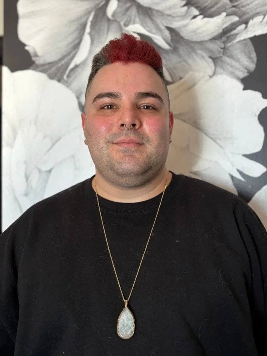 A person with short hair, partially dyed red on top, wearing a black shirt, a necklace with a large teardrop-shaped pendant, and a septum piercing, standing in front of a black and white floral background.