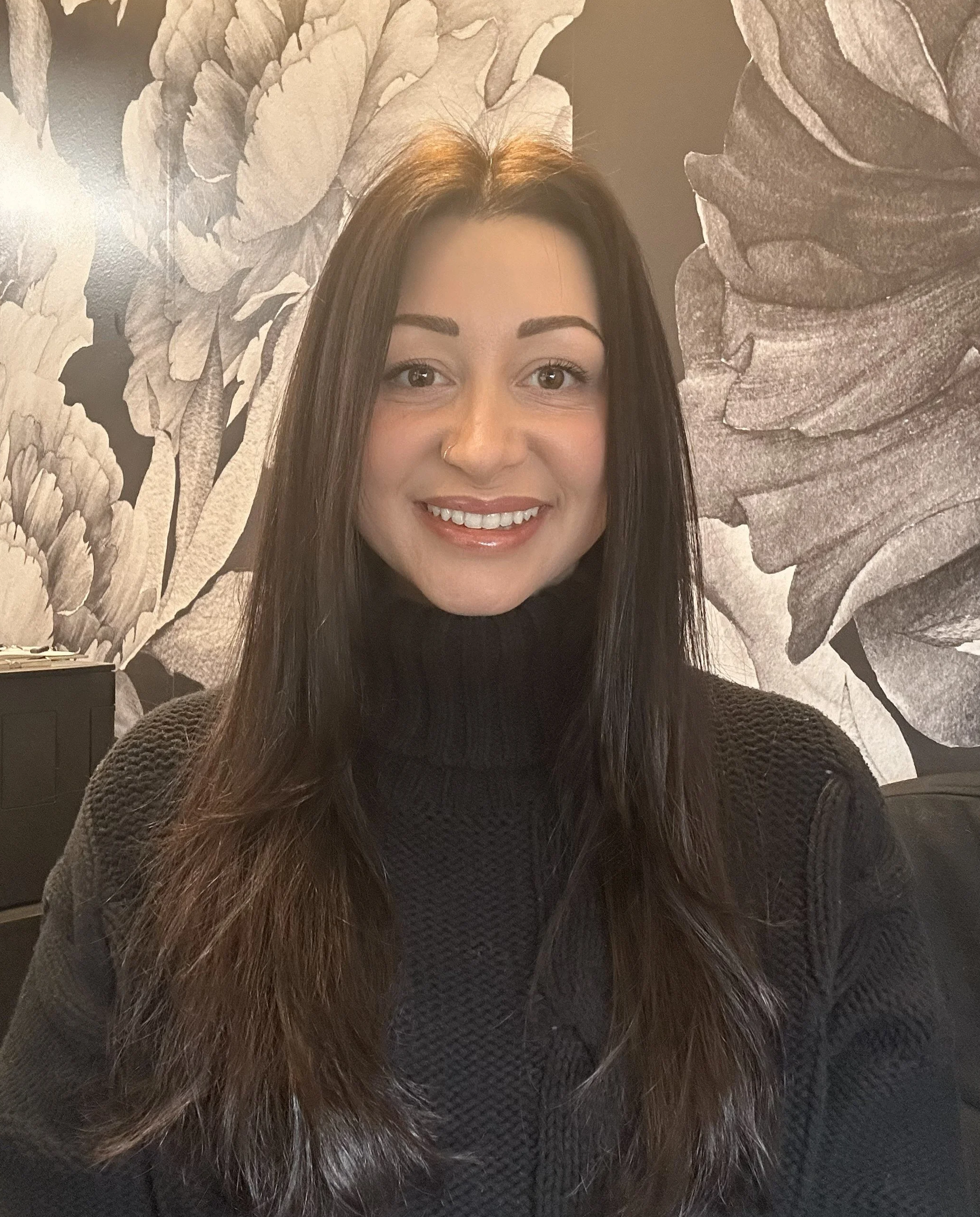 A smiling woman with long dark hair, wearing a black turtleneck sweater, seated indoors with a large floral mural on the wall behind her.