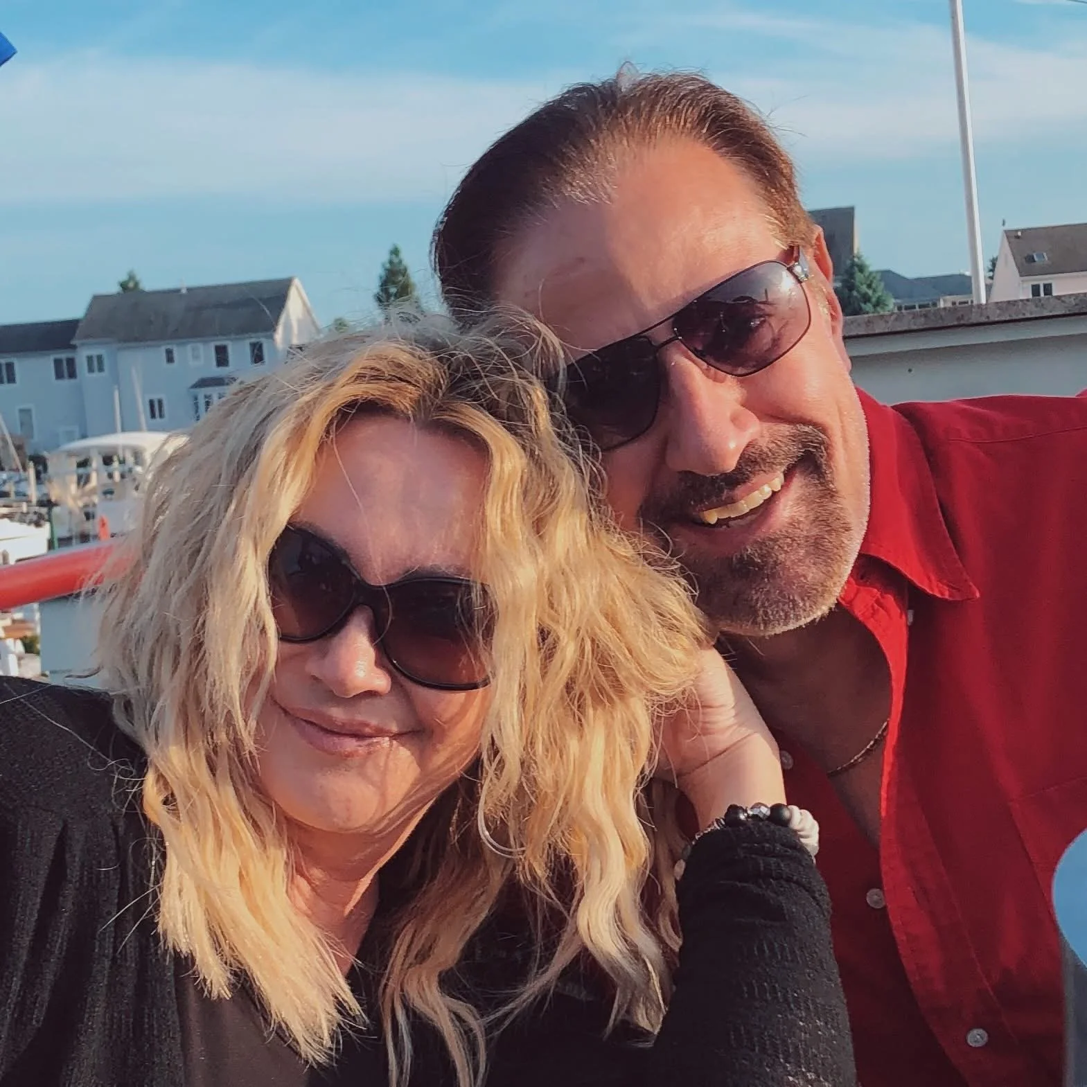 A smiling woman with curly blonde hair, sunglasses, and a black sweater, taking a selfie with a man wearing sunglasses, a red shirt, and a big smile, outdoors near a marina with boats and residential buildings in the background.
