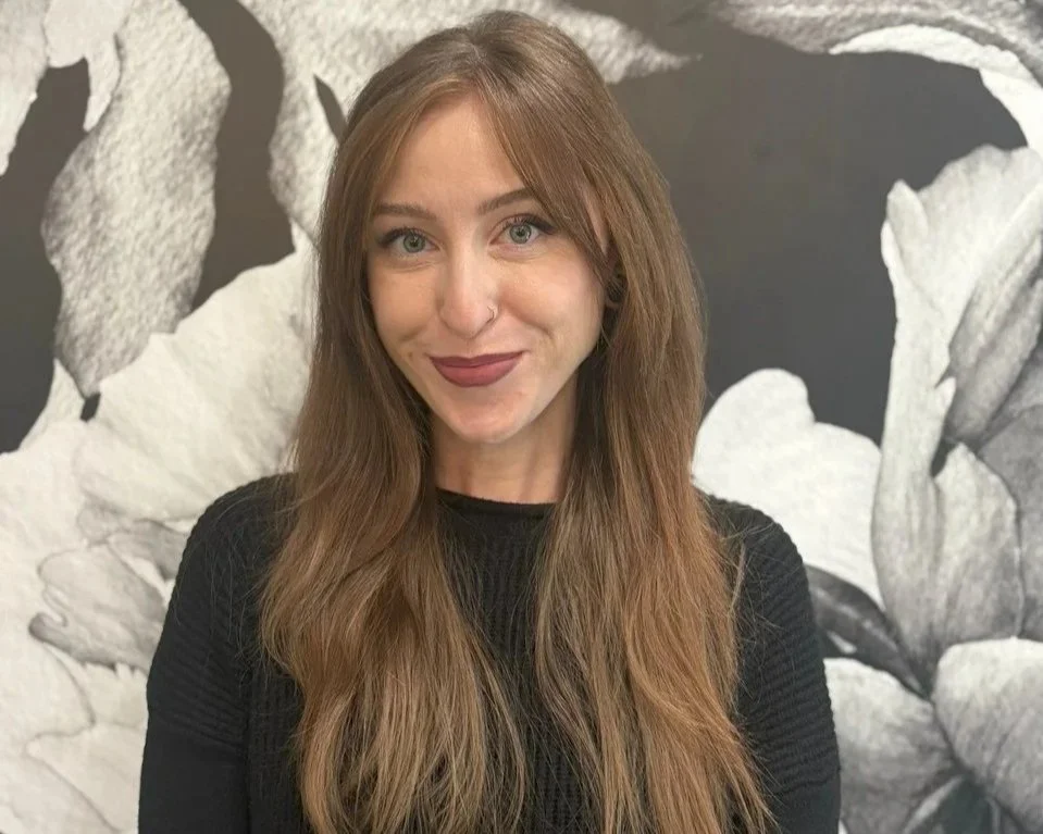 A woman with long brown hair and a nose piercing, smiling, wearing a black top, standing in front of a black and white floral mural.