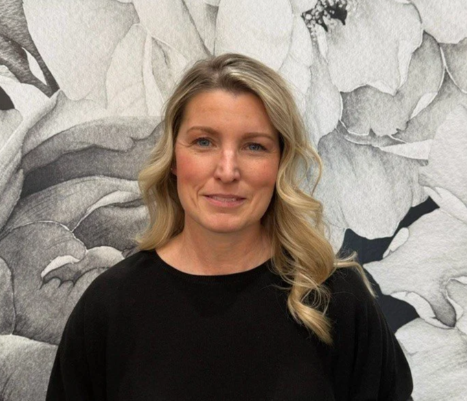 A woman with blonde, wavy hair wearing a black shirt standing in front of a black-and-white floral mural.