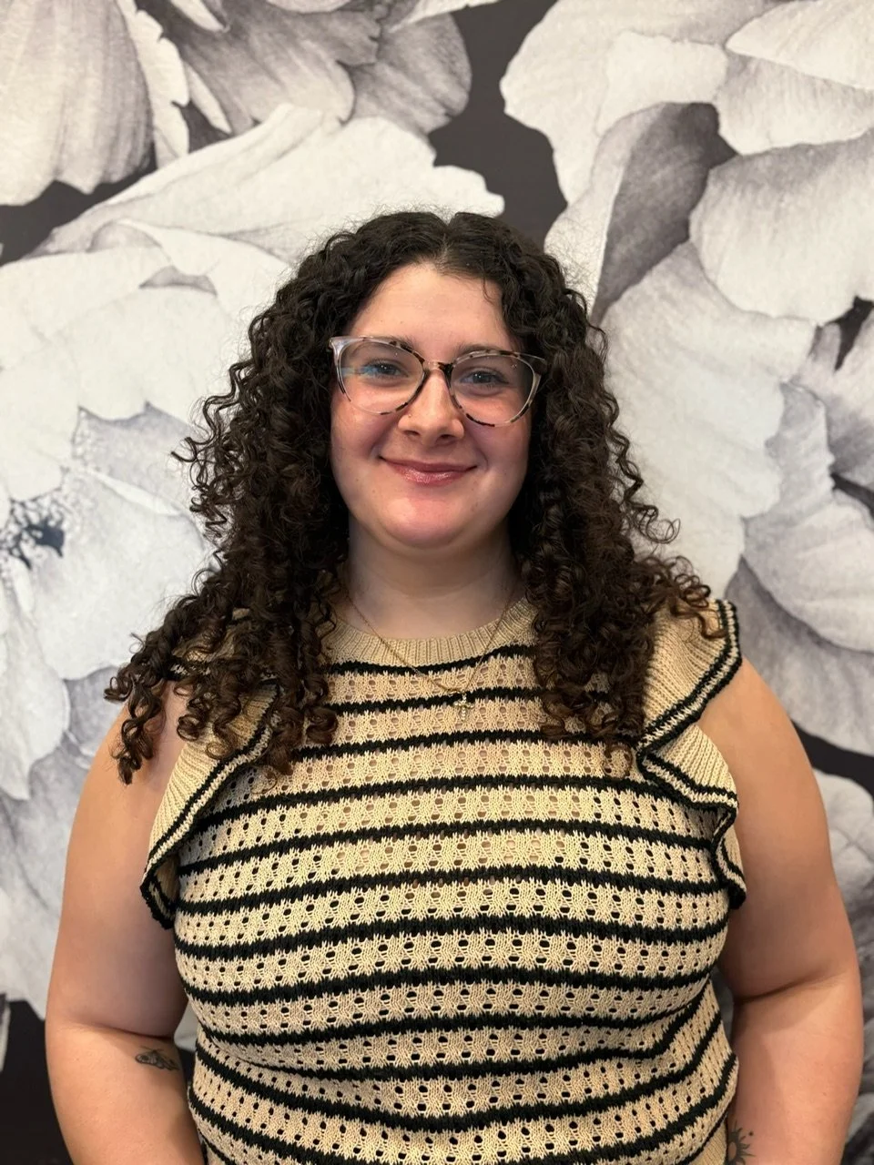 A woman with curly brown hair, glasses, and a light complexion smiling, standing in front of a black-and-white floral background.