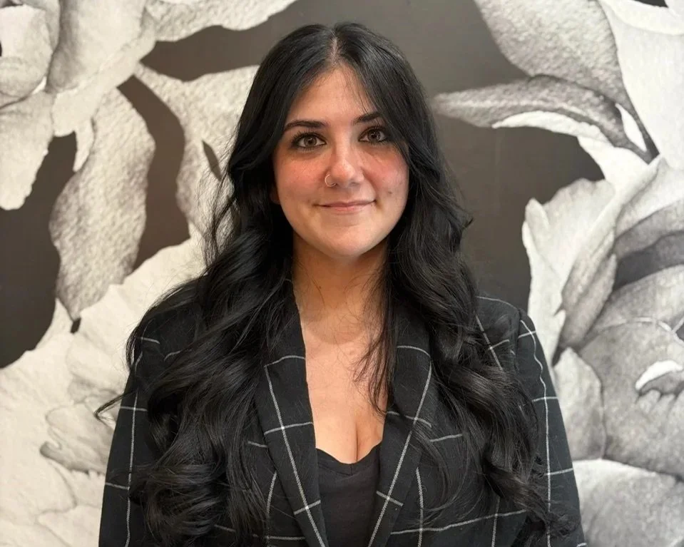A woman with long dark wavy hair, wearing a nose ring and a black blazer with white lines, standing in front of a black and white floral mural.