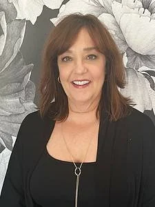 A woman with shoulder-length brown hair wearing a black top and a necklace, standing in front of a monochrome floral background.