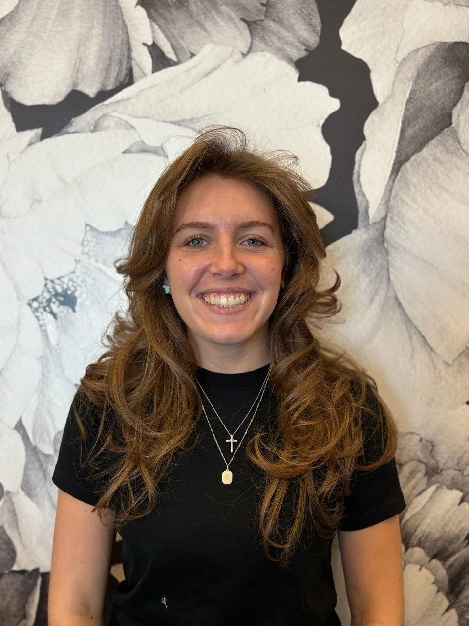 A young woman with long, wavy brown hair, smiling with teeth visible, wearing a black shirt and layered necklaces with crosses and initials, standing in front of a black-and-white floral mural.