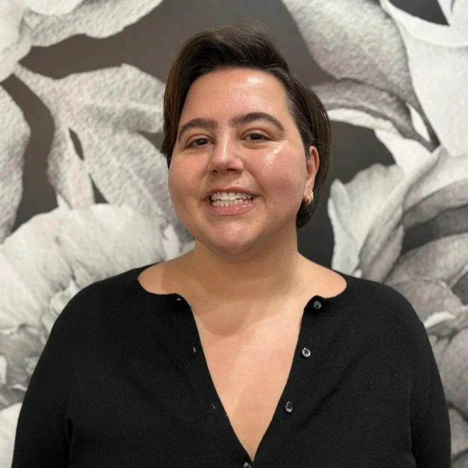 A woman with short dark hair, wearing a black button-up top, smiling and standing in front of a black and white floral background.