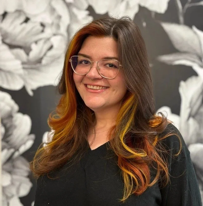 A woman with long, wavy hair featuring shades of red, orange, and yellow, wearing glasses and a black top, smiling in front of a black-and-white floral patterned background.