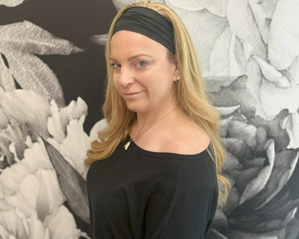 A woman with long blonde hair wearing a black headband and a black top standing in front of a black and white floral mural.