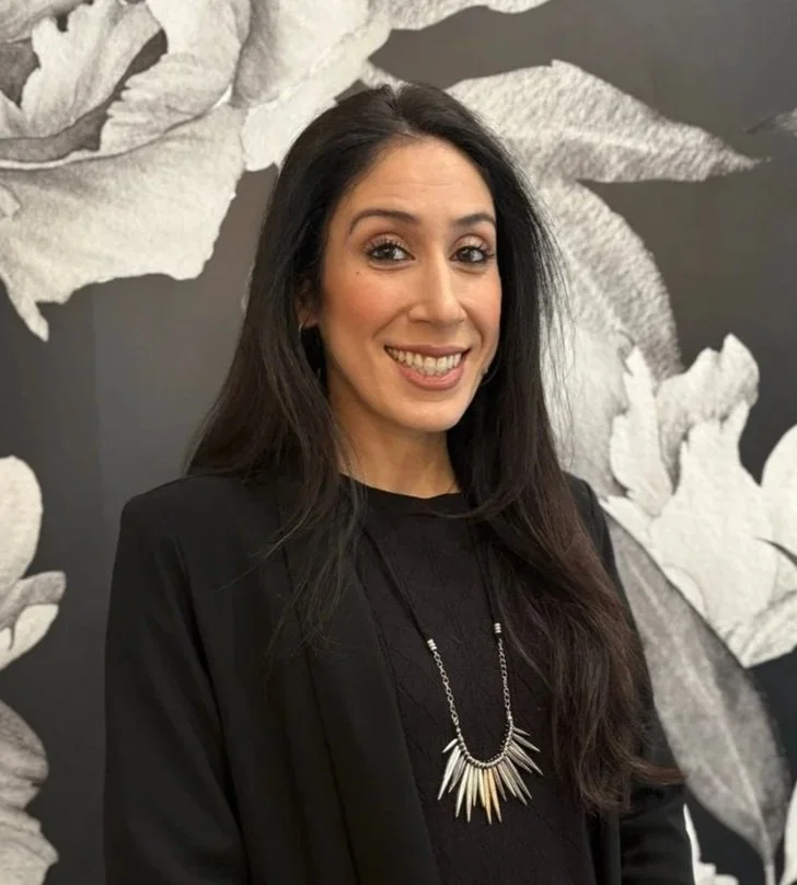 A woman with long dark hair, smiling, dressed in business attire, standing in front of a large gray and white floral wall art.