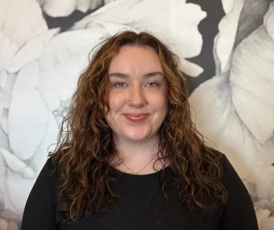 A woman with curly, shoulder-length brown hair, wearing a black top, standing in front of a black and white floral mural background.