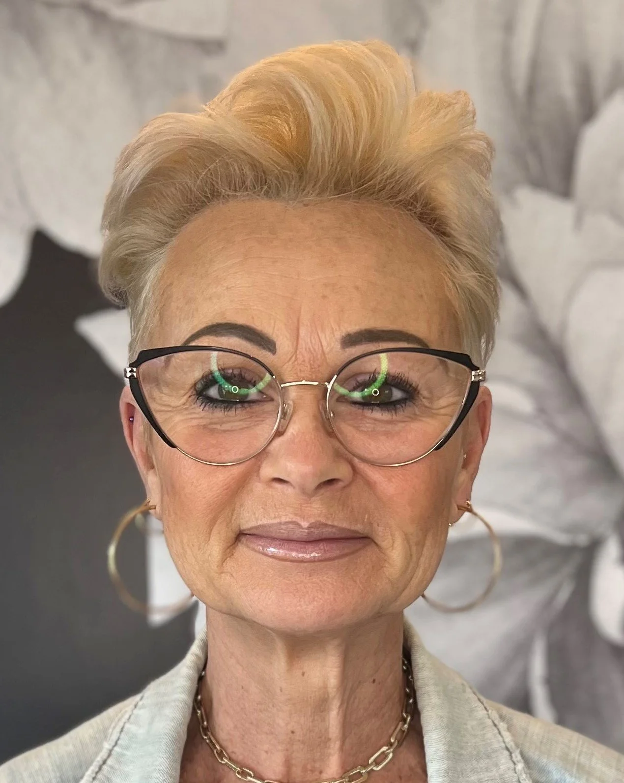 Close-up of an older woman with short blond hair, glasses, and gold hoop earrings, smiling at the camera.