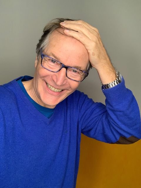 Ein lachender älterer Mann mit Brille, der die Hand auf den Kopf legt und ein blaues Hemd trägt.