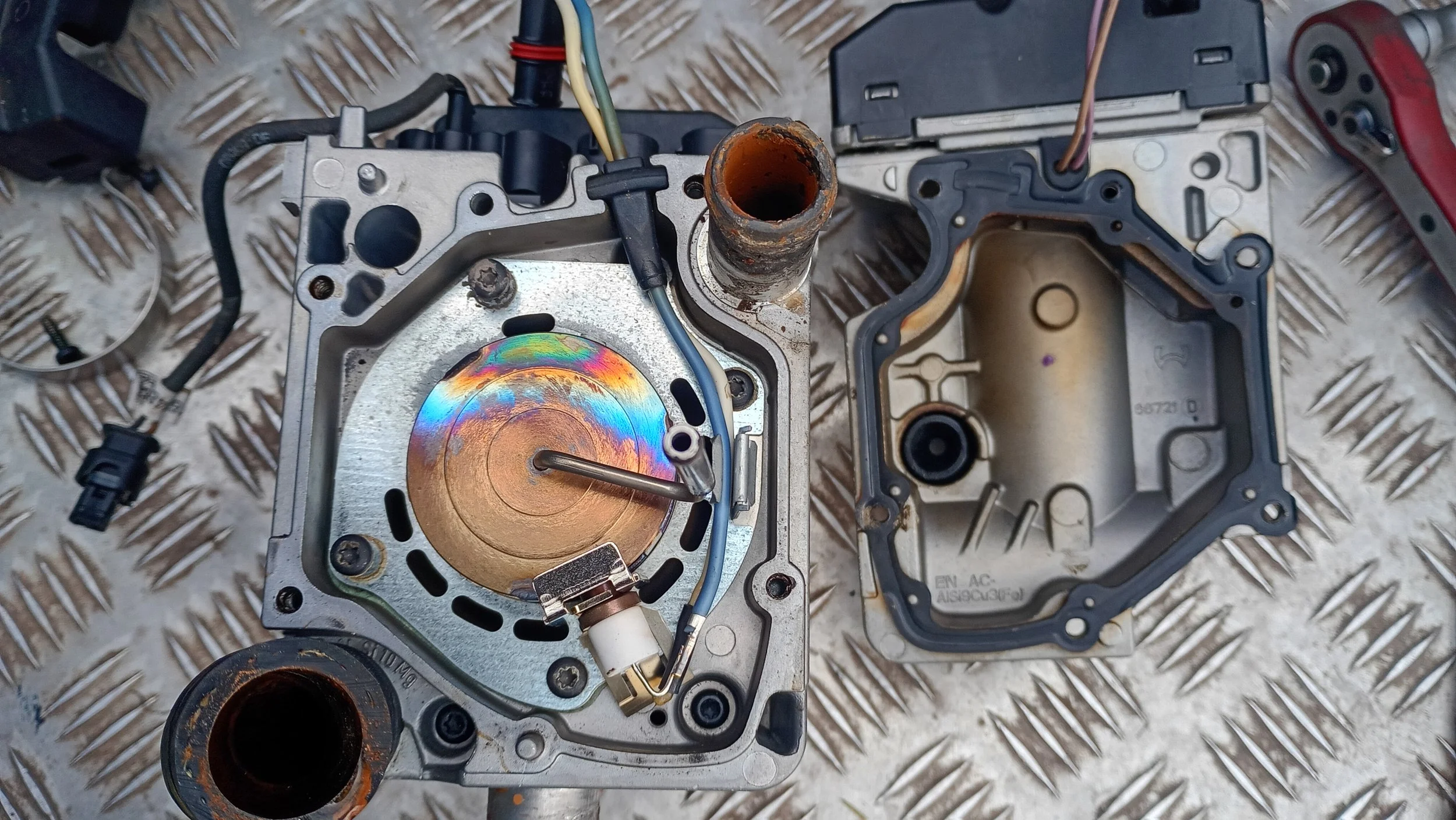 Disassembled mechanical device with metal parts, wiring, and rusted openings, placed on a checkered metal surface.