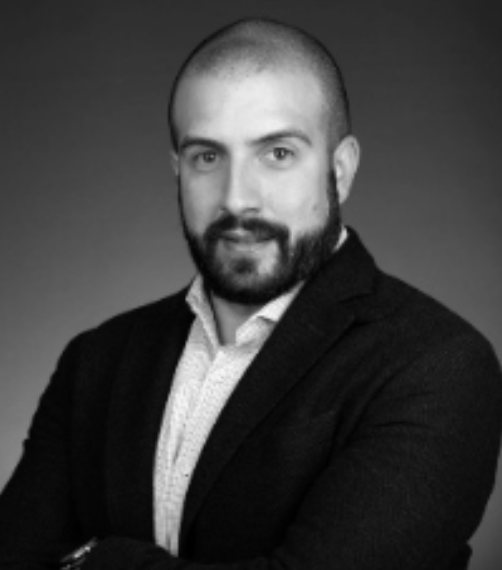 Black and white professional headshot of a man with a beard, short hair, wearing a suit jacket and a collared shirt.