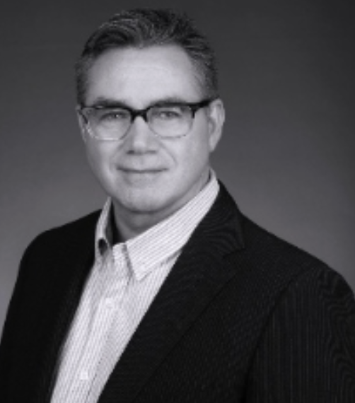 Black and white portrait of a man wearing glasses, a light-colored dress shirt, and a dark blazer, smiling slightly.