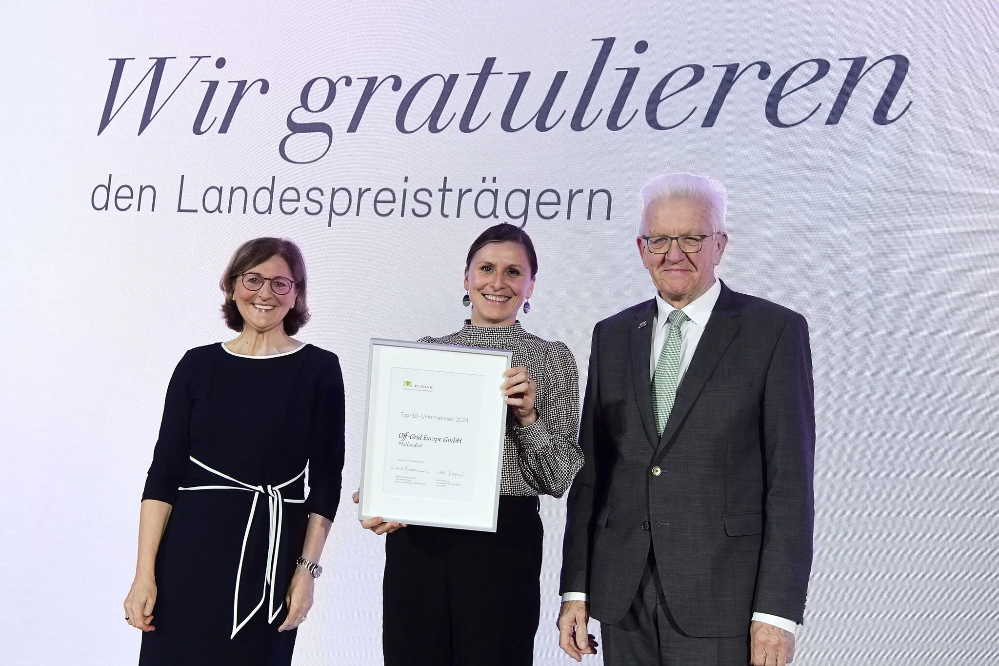 Three people standing on a stage, smiling. The woman in the middle is holding a framed award. The background has large text in German that says, 'Wir gratulieren den Landespreis Trägern'.