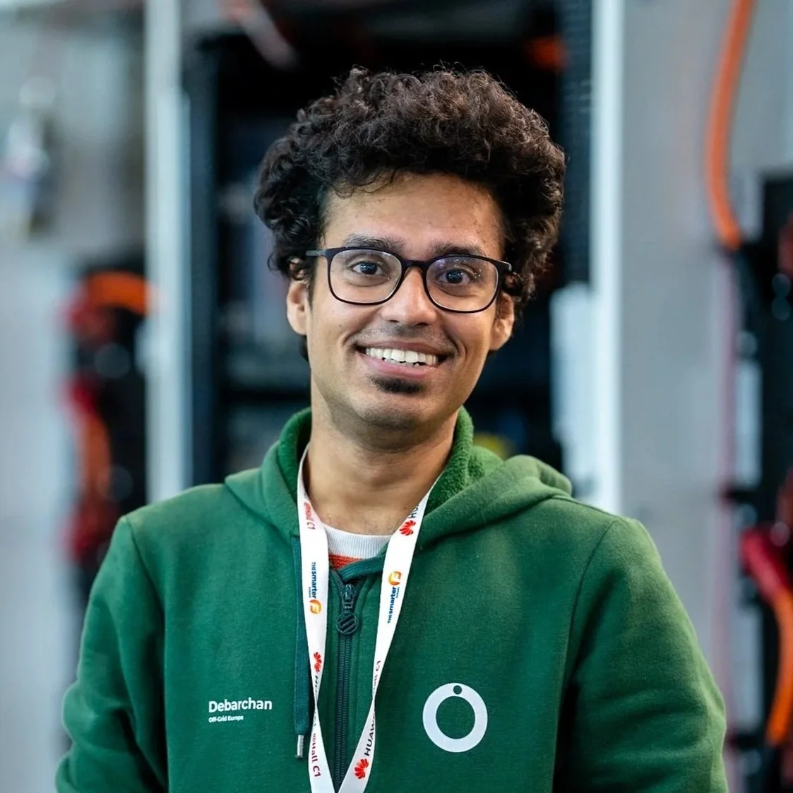 A smiling man with glasses, curly dark hair, wearing a green hoodie with a logo and name tag, standing in front of server racks or computer equipment.
