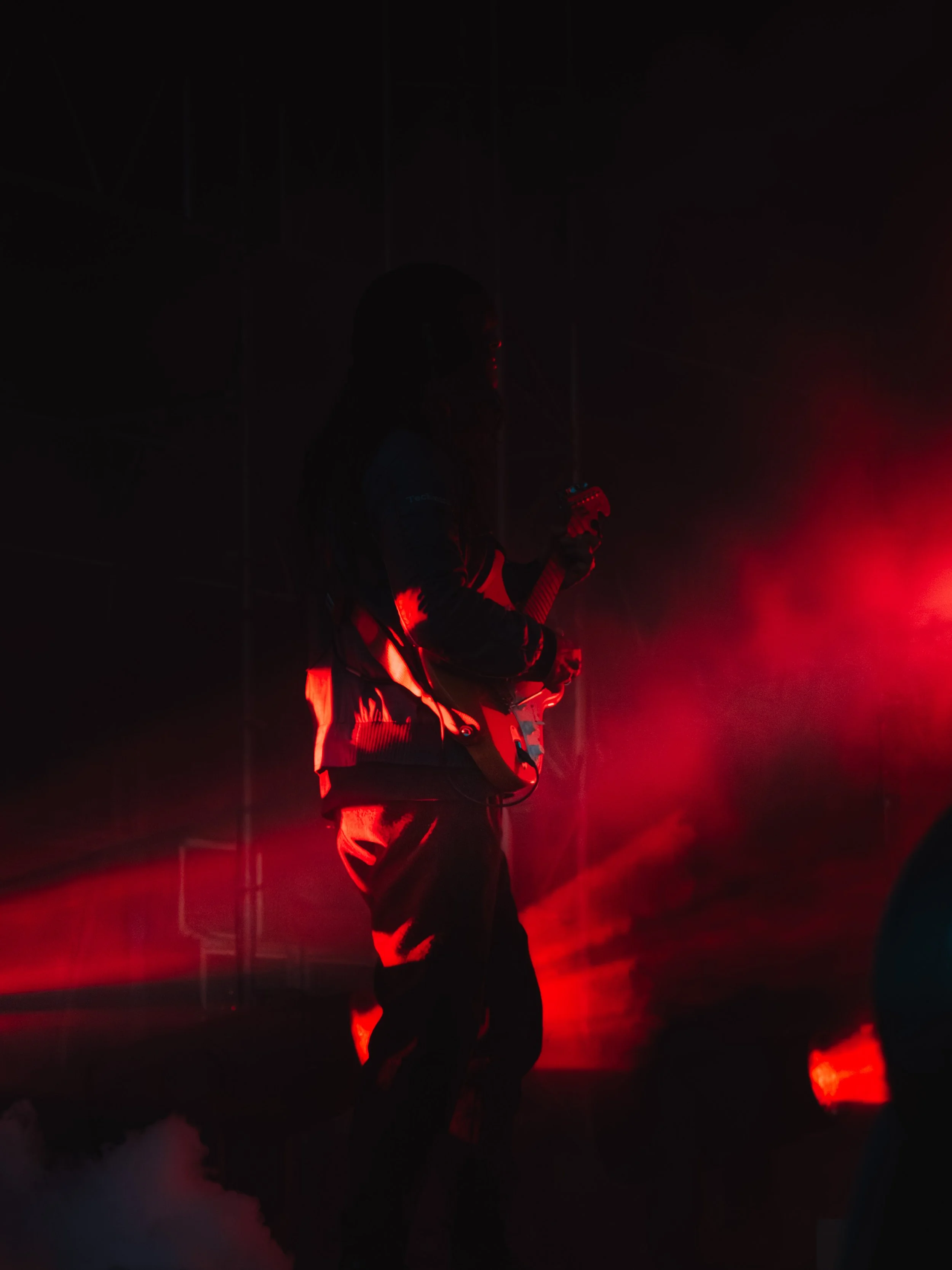 Person playing an electric guitar on stage at night, illuminated by red stage lighting, with fog effects.