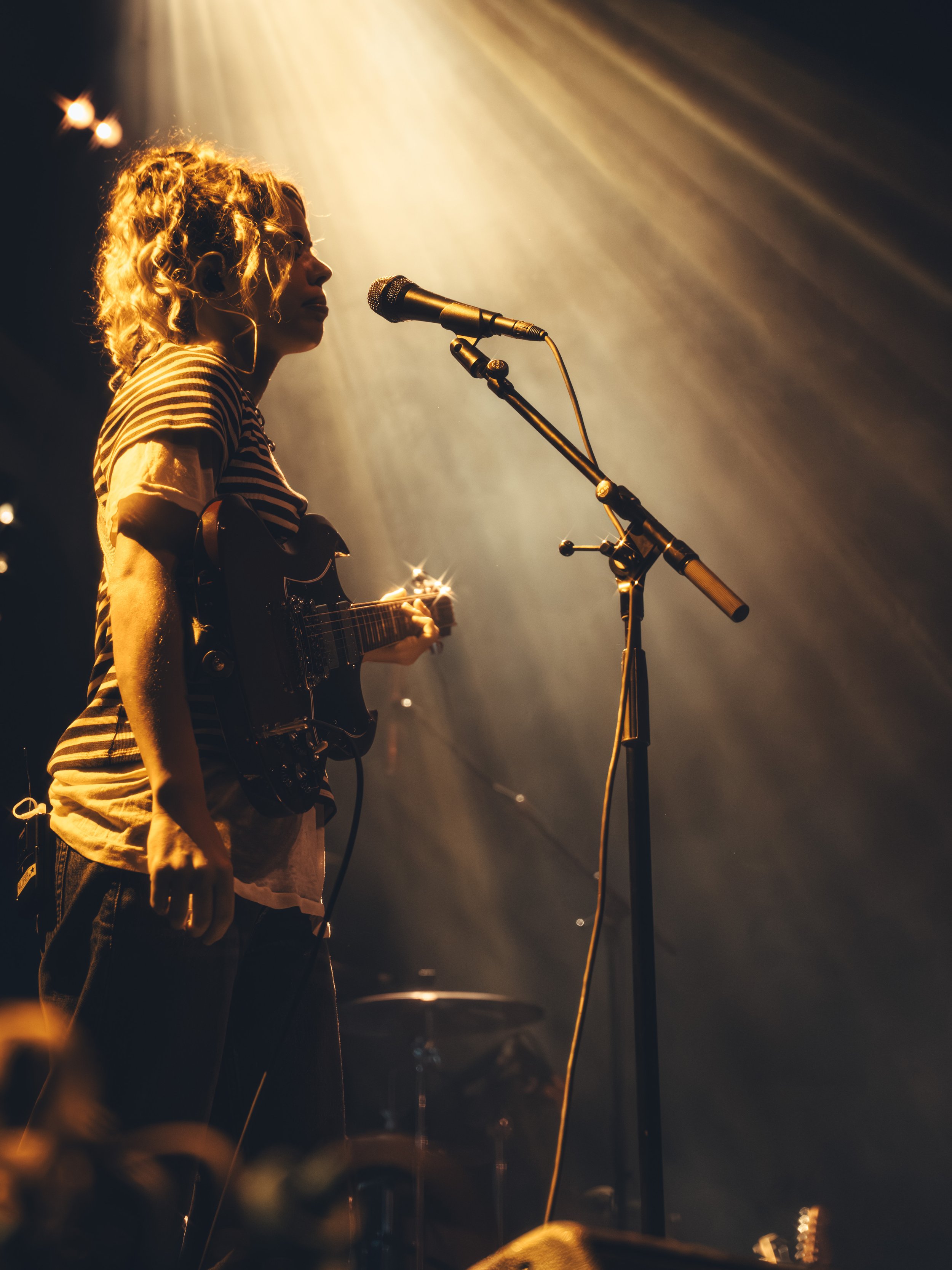 A female musician with curly blonde hair playing an electric guitar on stage, standing in front of a microphone with stage lighting creating a warm glow.